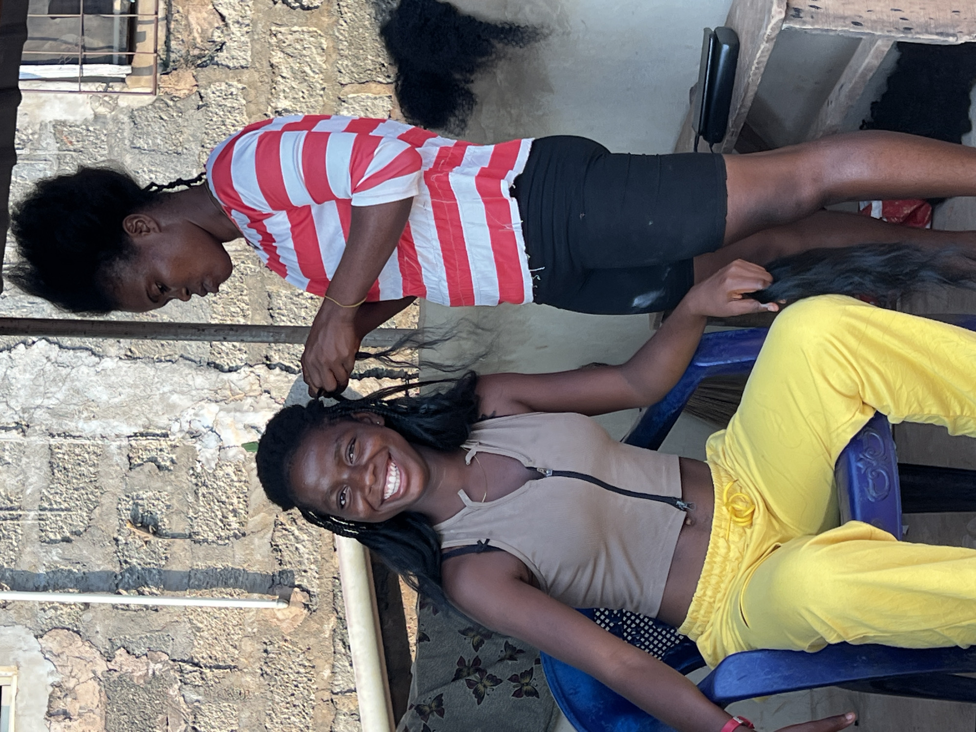 The image shows two young Black women in an outdoor setting, likely a residential area in Nkpor, Nigeria. One woman, wearing a red and white striped t-shirt and black shorts, is standing and braiding the hair of the other woman. The second woman is seated on a blue plastic chair and is wearing a tan zippered top and bright yellow pants. She is smiling at the camera, appearing happy and relaxed. The background shows a rough, textured stone wall and a wooden table, suggesting a somewhat rustic environment. The lighting indicates it is daytime. The activity is hair braiding, a common practice in many cultures.