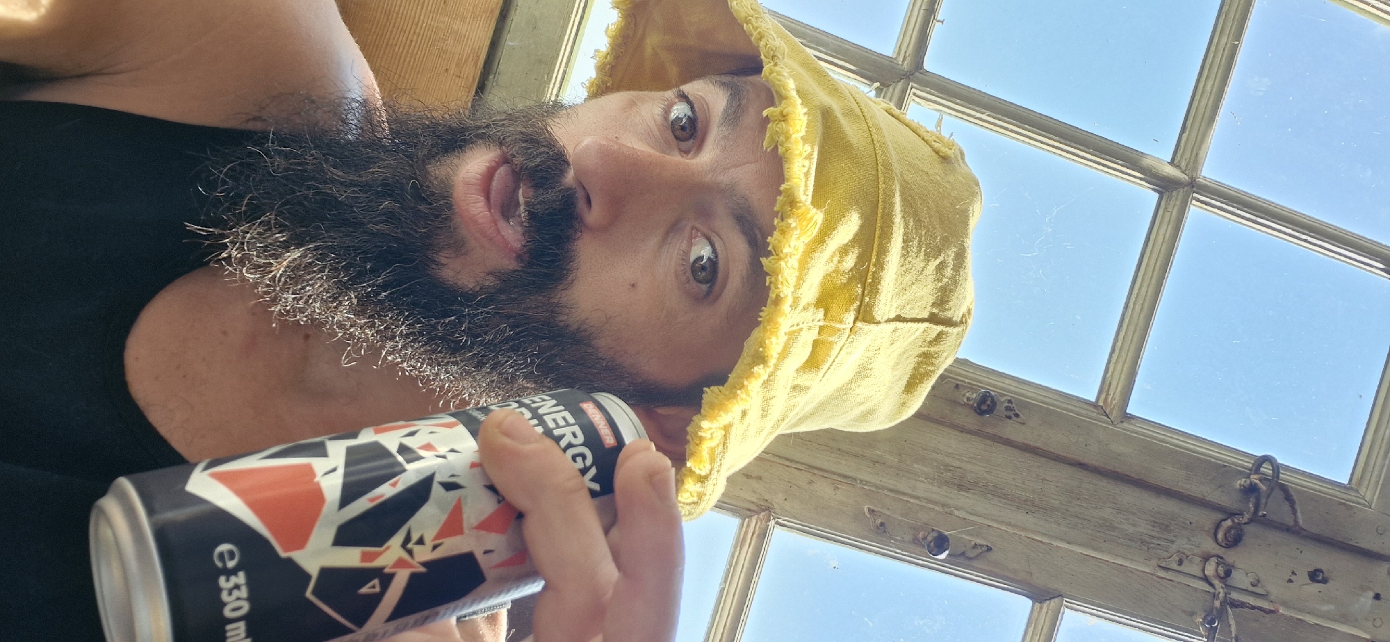 The image is a close-up selfie of a man wearing a yellow bucket hat and a black tank top. He has a beard and is holding a can of energy drink. The background shows a window with a view of the sky. The man is likely indoors, possibly in Wattenwil, Switzerland, given the context.
