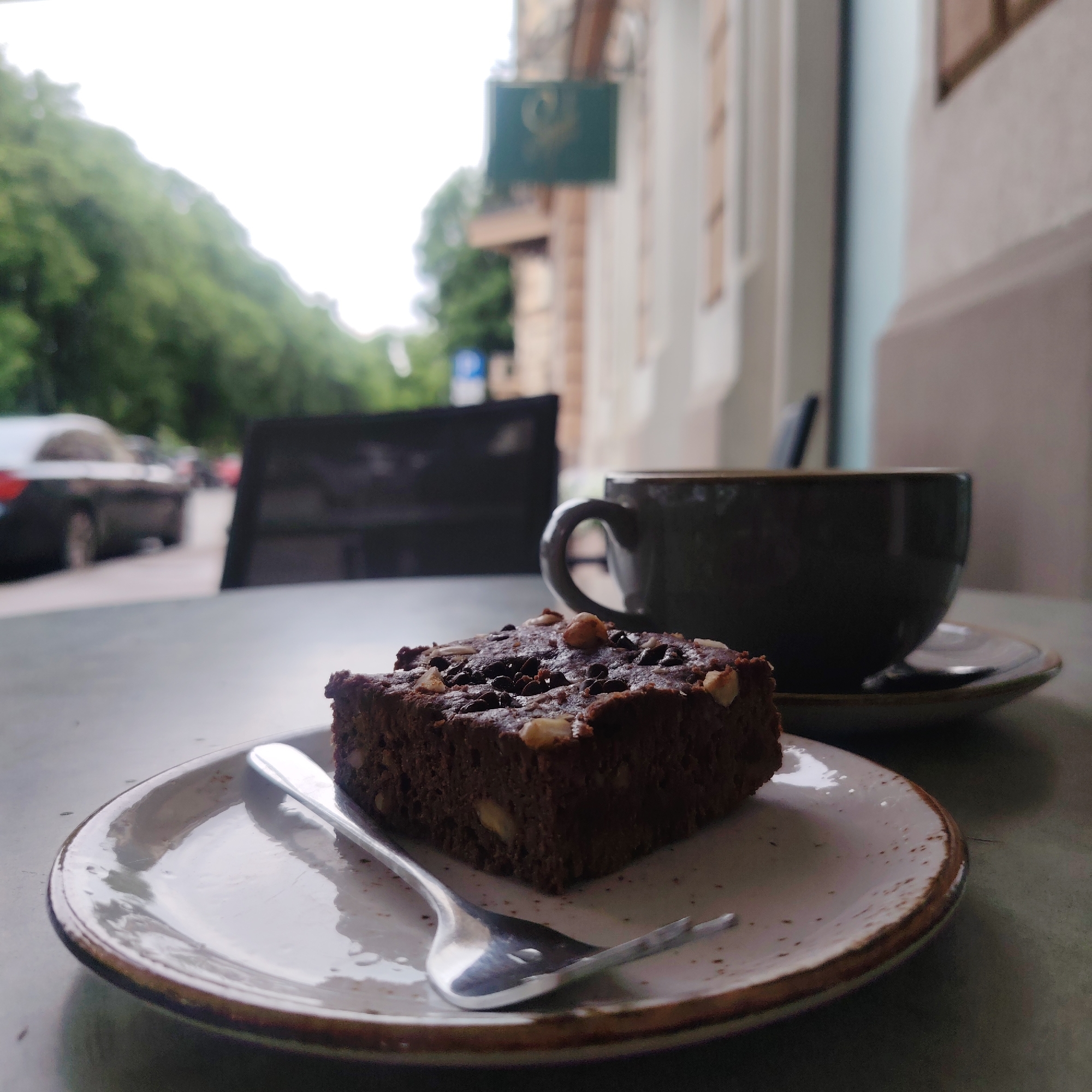 A close-up, eye-level shot captures a brownie and a dark grey mug of coffee on a table outside a building. The brownie, rich and dark chocolate, sits on a speckled white plate with a rustic brown rim. It's topped with what appear to be chocolate chips and chopped nuts. A silver fork rests on the plate, with its tines partially inserted into the brownie. To the right, a dark grey mug with a thick handle sits on a matching saucer, containing coffee.
In the background, the scene is slightly blurred, hinting at an urban setting. Trees with green foliage line a street, and a dark-colored car is visible in the distance. A building with windows and a sign is also present. The overall lighting suggests an overcast day. The location is identified as Riga, Latvia.