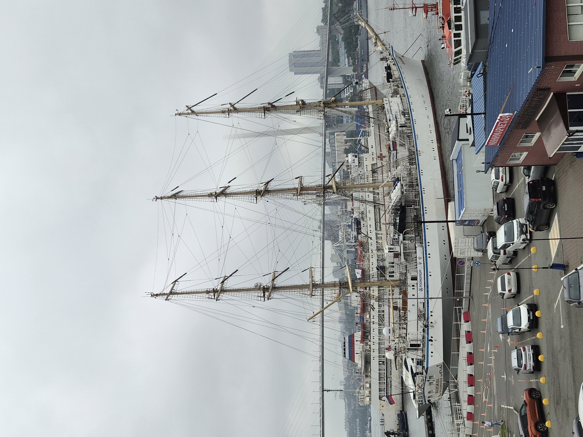 The image shows a large white sailing ship with three masts docked in a harbor in Vladivostok, Russia. The ship has the name "НАДЕЖДА" and "ВЛАДИВОСТОК" written on its hull. In the background, a large cable-stayed bridge spans the water, with a cityscape visible on the other side. There is a parking lot in the foreground with several cars parked. A few people are visible walking around the parking lot and on the ship. The sky is overcast. The building in the foreground has a sign that reads "МИНИМАРКЕТ 24".