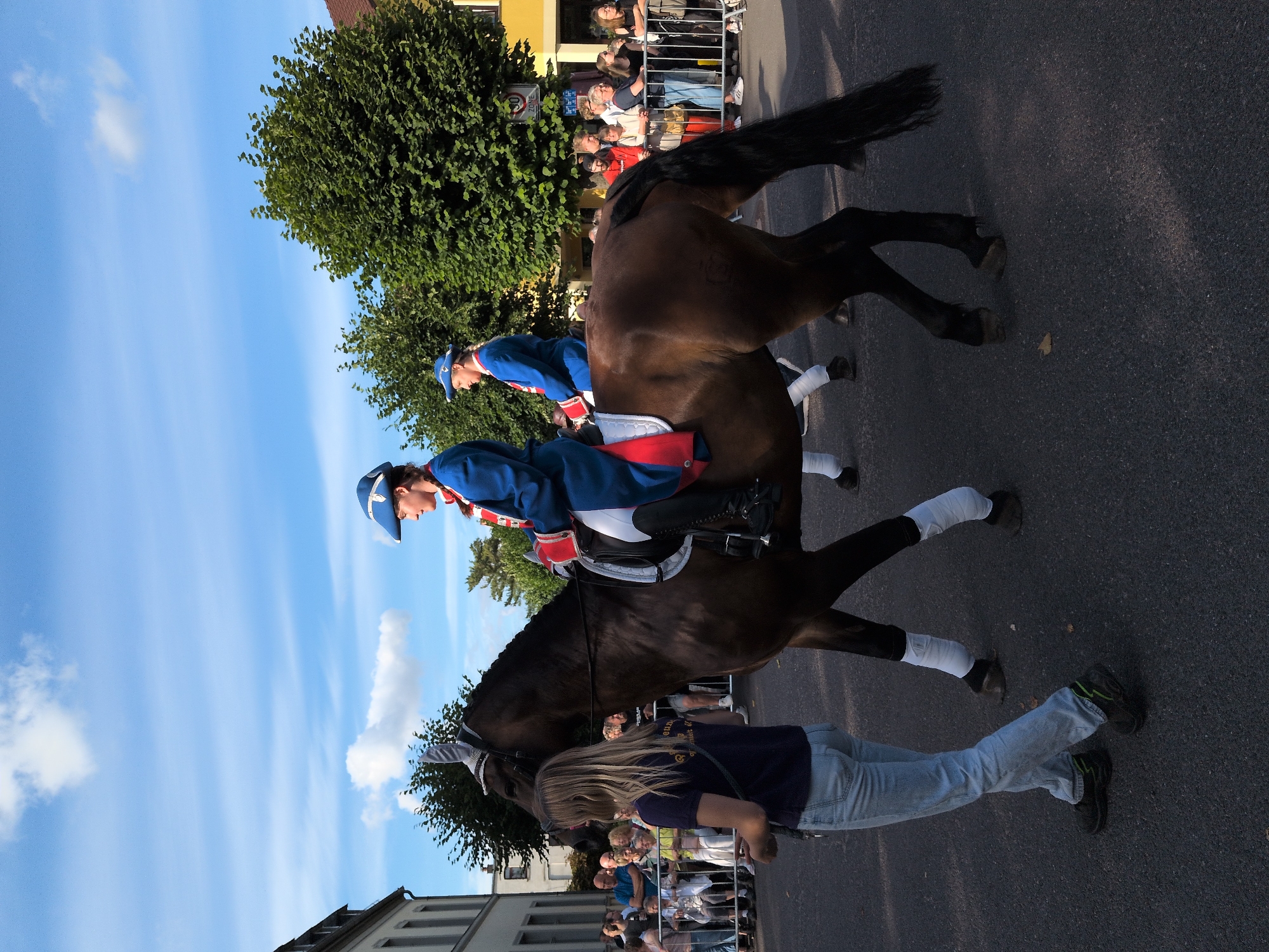 A parade is taking place on a street in Buttstädt, Germany. The sky is blue with some white clouds. On the right side of the image, a yellow building with a tiled roof is visible. In the background, there are trees. In the foreground, a dark brown horse is walking from left to right, ridden by two individuals in blue historical uniforms with white and red trim. The rider in front is wearing a blue hat with a silver emblem, a blue jacket with red cuffs and lapels, and black boots. The rider behind is wearing a similar uniform and hat. A woman with blonde hair, wearing a dark blue t-shirt and light blue jeans, is walking next to the horse on the left, holding a leash. Spectators are lined up behind a fence on the left and right sides of the street, watching the parade. The street surface is asphalt.