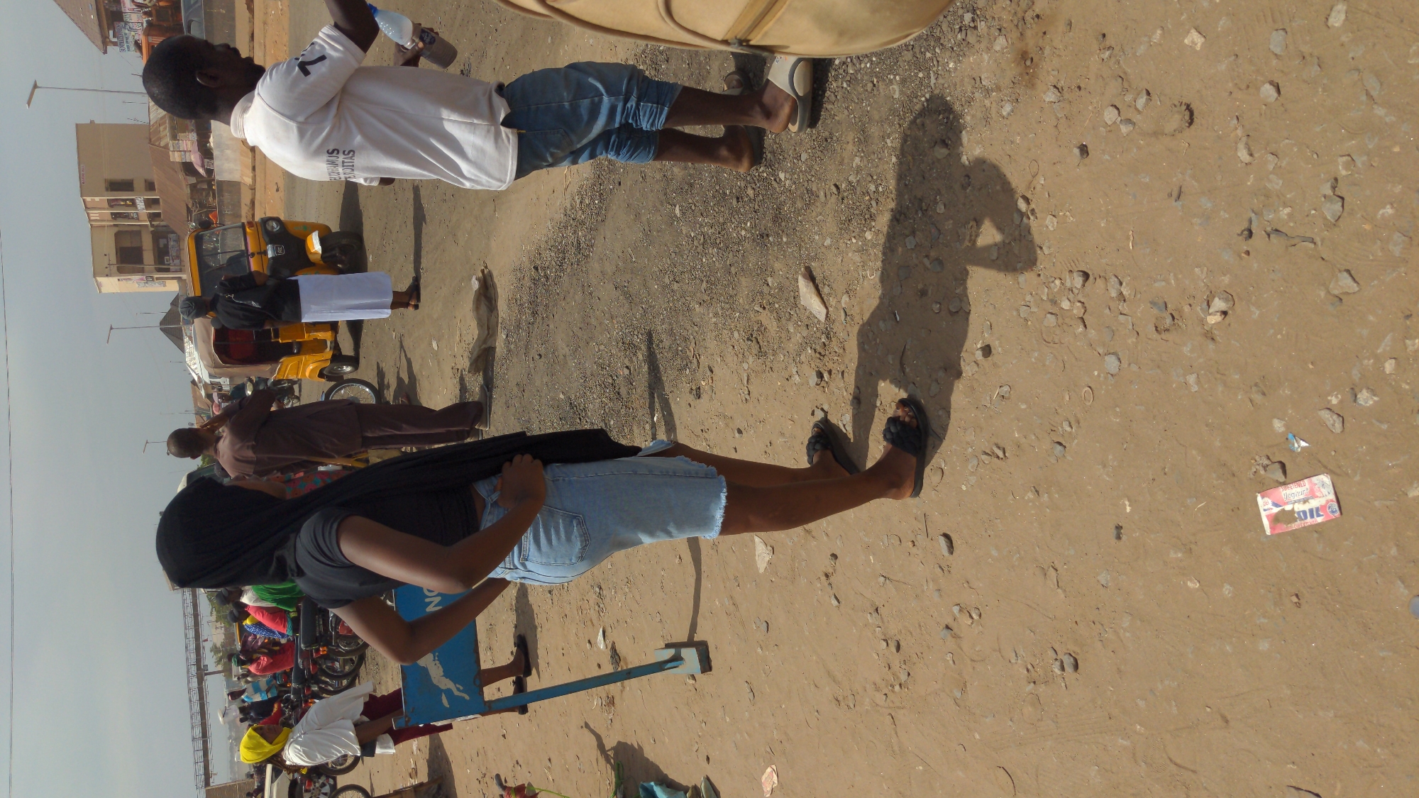 The image captures a street scene in Ado, Nigeria. The ground is sandy and gravelly, with scattered rocks and debris, including a discarded box of oil. Two people are prominent: one wearing a white shirt and blue shorts, holding a water bottle, and another with a black hijab, dark gray shirt, and blue jean shorts. The background shows a yellow tricycle, several parked motorcycles, and other individuals. Buildings are seen in the distant background under a bright sky.