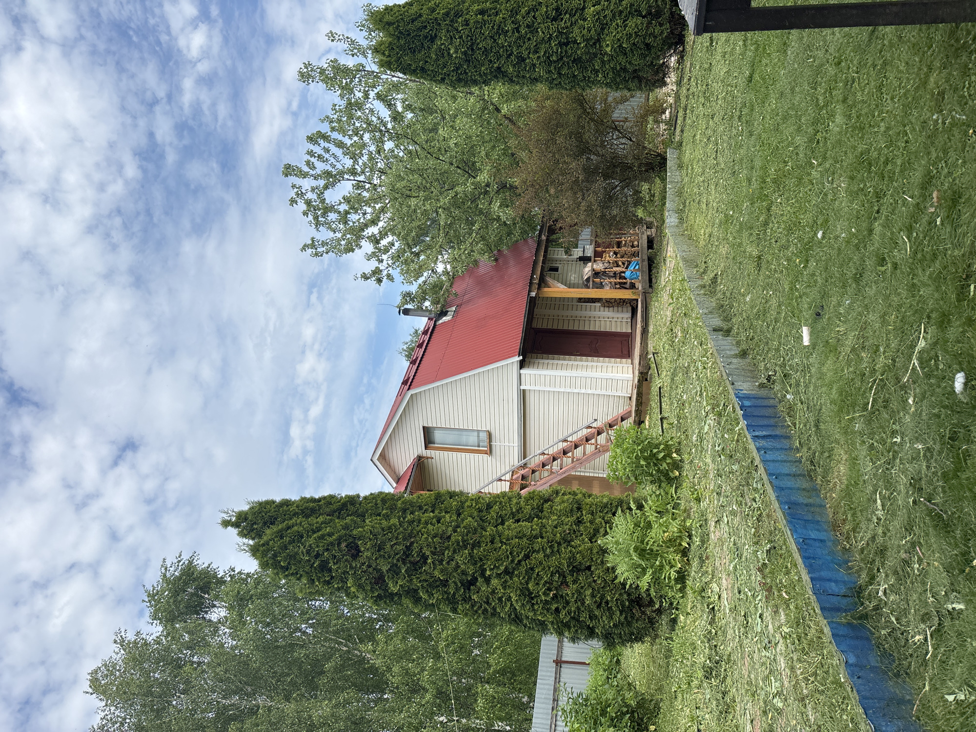 The image depicts a single-story house in what appears to be a rural or suburban setting. The house has a light-colored siding and a red roof, with a wooden staircase leading to a door. A chimney is visible on the roof. The house is surrounded by greenery, including tall coniferous trees, deciduous trees, and grass. A blue fence runs along the right side of the image. The sky is partly cloudy. The image does not provide enough information to confirm that the location is in the city of Мещёры, Russia. There are no people visible in the image.