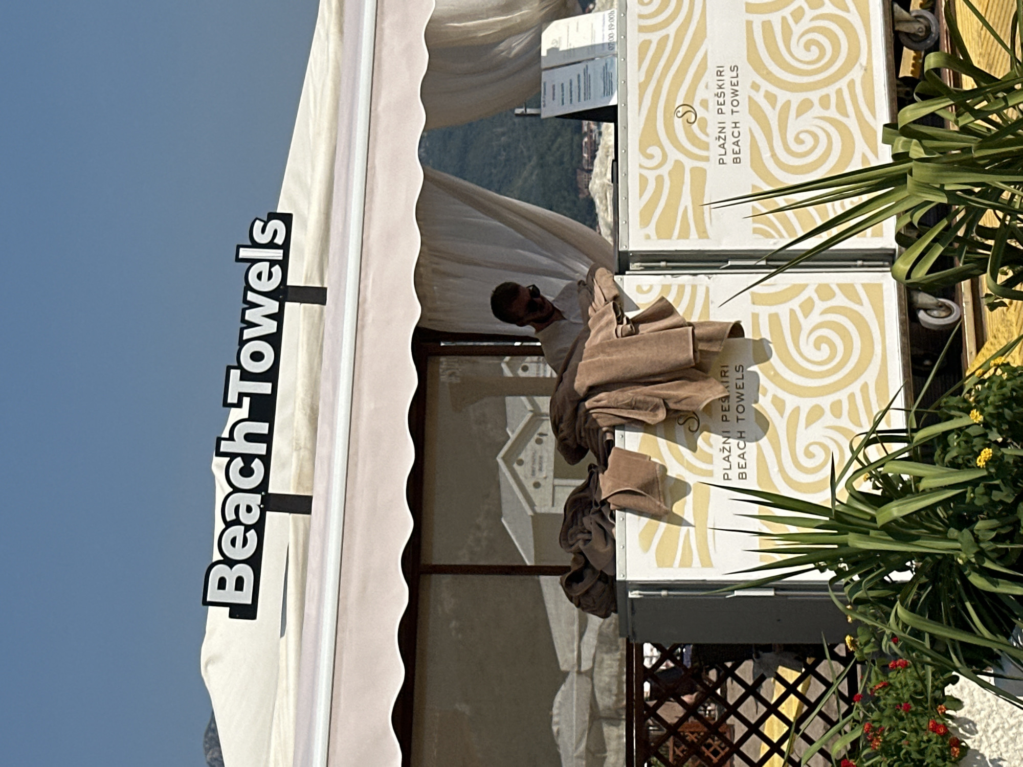 This is a photograph of a beach towel rental stand in Boreti, Montenegro. The stand has a white awning with scalloped edges and a sign reading "Beach Towels." A man wearing sunglasses and a white shirt stands behind the counter, looking down. Several brown beach towels are piled on the counter. The stand is decorated with a yellow wave pattern. In the background, there are buildings, mountains, and a clear blue sky. The scene appears to be set up for a sunny day at the beach.