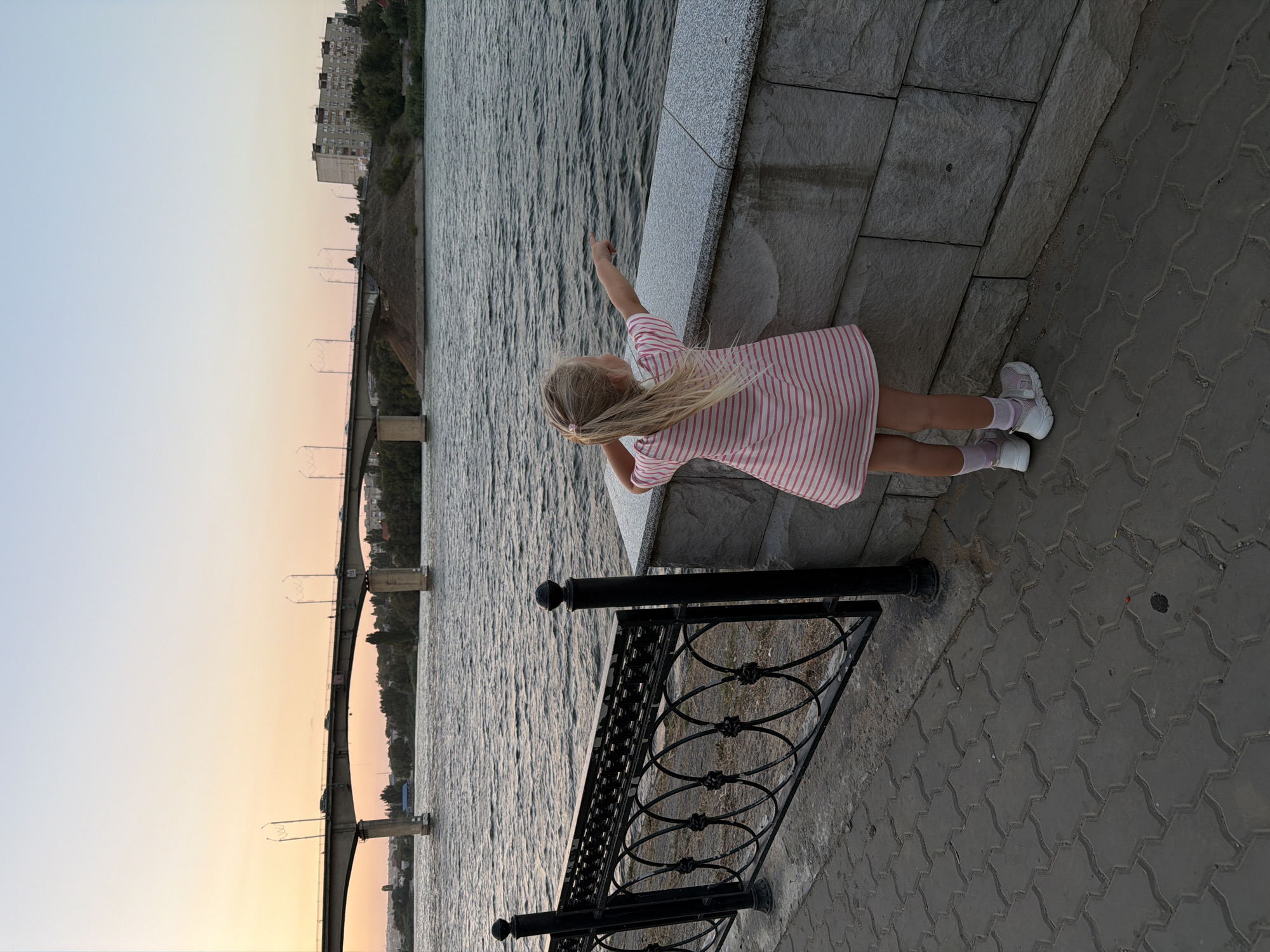 A young girl with blonde hair, wearing a pink and white striped dress and white sneakers, stands facing away from the camera, pointing towards the Volga River. She is positioned on a paved walkway next to a decorative black metal fence and a stone wall. In the background, a wide river stretches towards a bridge with multiple arches and traffic. The sky above is a soft gradient of pale orange and blue, indicating either sunrise or sunset. Along the far bank of the river, trees and buildings are visible, suggesting an urban or semi-urban setting. The location is identified as the embankment of the Volga River in Kamyshin, Russia.