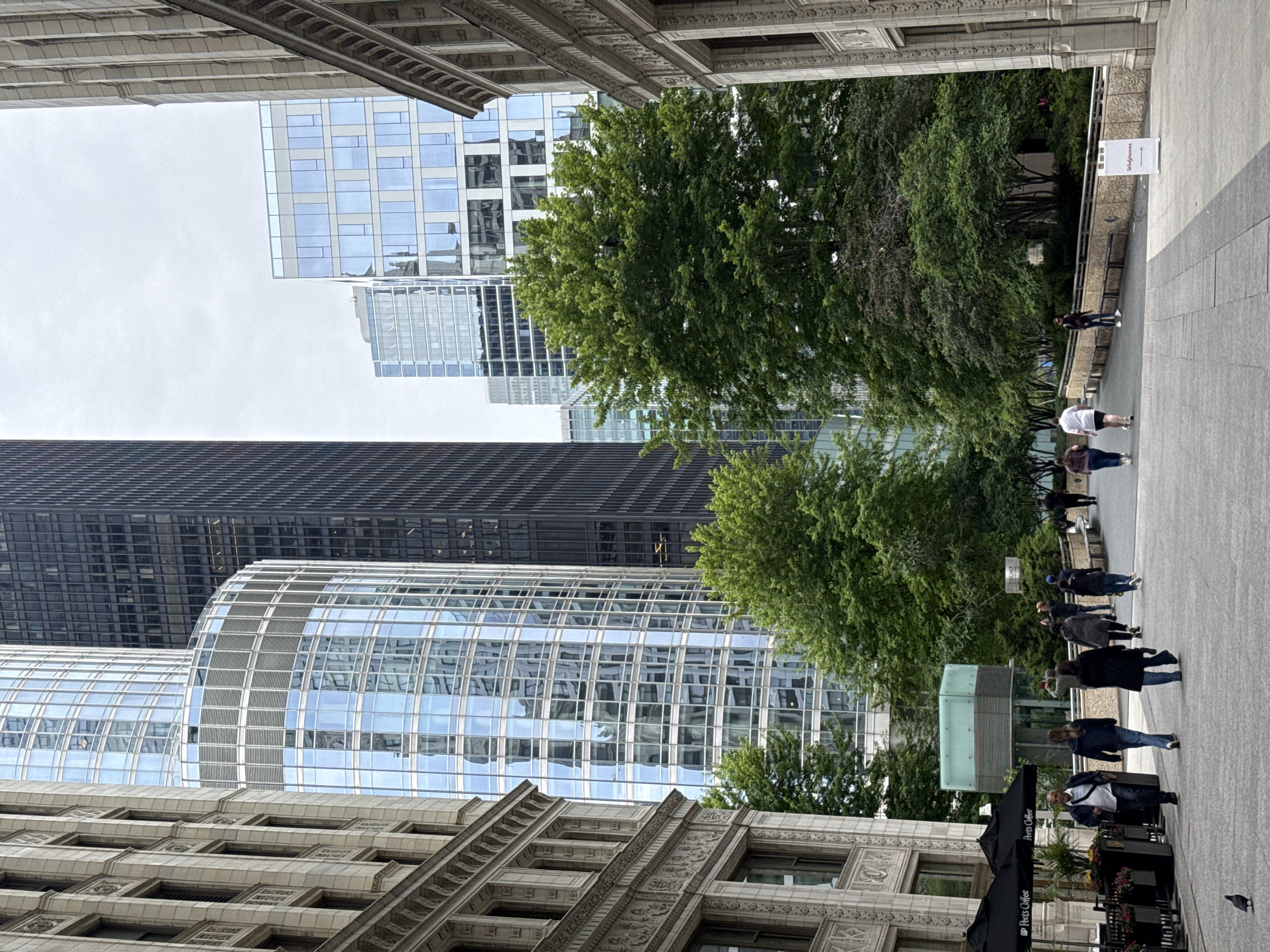 In Chicago, Illinois, USA, a street scene unfolds. Tall buildings, a mix of modern glass structures and older stone edifices, dominate the background. Lush green trees punctuate the urban landscape, with their foliage providing a natural contrast to the architecture. Several people are visible walking along a paved walkway, suggesting a public space within the city. The sky appears overcast, casting a diffused light over the scene. One of the buildings displays the "Peet's Coffee" logo, indicating a commercial establishment. A pigeon is present on the walkway, adding a small detail of urban wildlife. The overall impression is one of a bustling metropolitan area with a blend of natural and man-made elements.