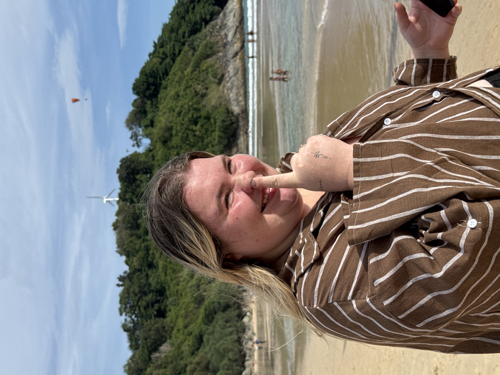 A woman with blonde hair is shown at a beach, smiling and touching her nose with her thumb. She is wearing a brown and white striped shirt. In the background, there is the ocean with waves, and a green, tree-covered hill. A windmill and a paraglider are visible in the distance. The beach appears to be located in Rawai, Thailand.