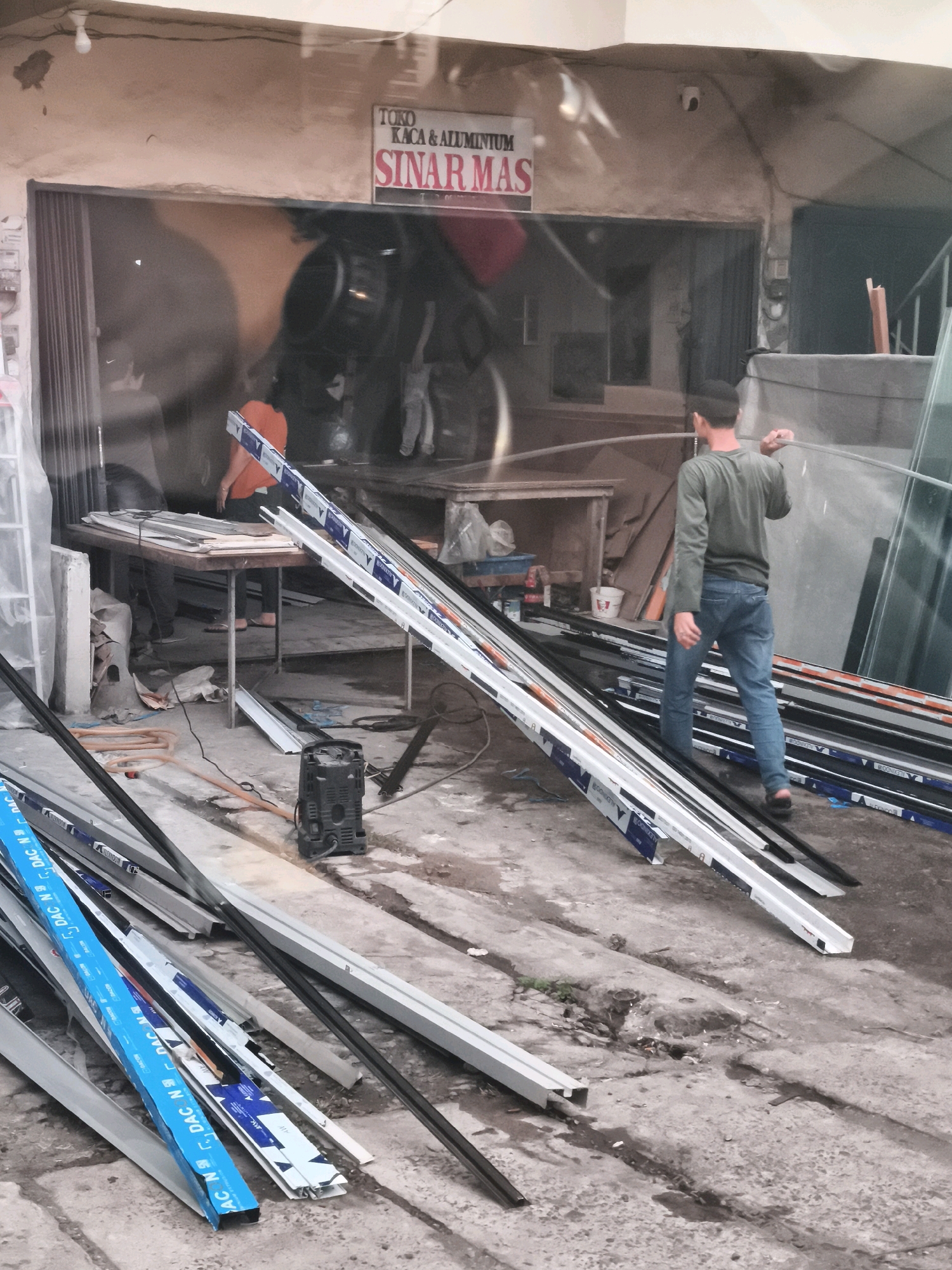 The image depicts an outdoor scene in front of a shop with a sign reading "TOKO KACA & ALUMINIUM SINAR MAS" in Palembang, Indonesia. The shop appears to sell glass and aluminum products, as indicated by the various aluminum profiles and glass panes visible. A man in a green long-sleeved shirt and blue jeans is walking away from the viewer, carrying a long, clear object, possibly a glass sheet or plastic tubing. Another person, wearing an orange shirt, is partially visible inside the shop entrance, standing near a table laden with aluminum extrusions. The ground is concrete and shows signs of wear and tear, with scattered debris. In the foreground, a pile of aluminum profiles is stacked, some with blue and white branding. A black industrial fan is present on the ground. The overall impression is one of a busy workshop or storage area associated with a construction materials business.