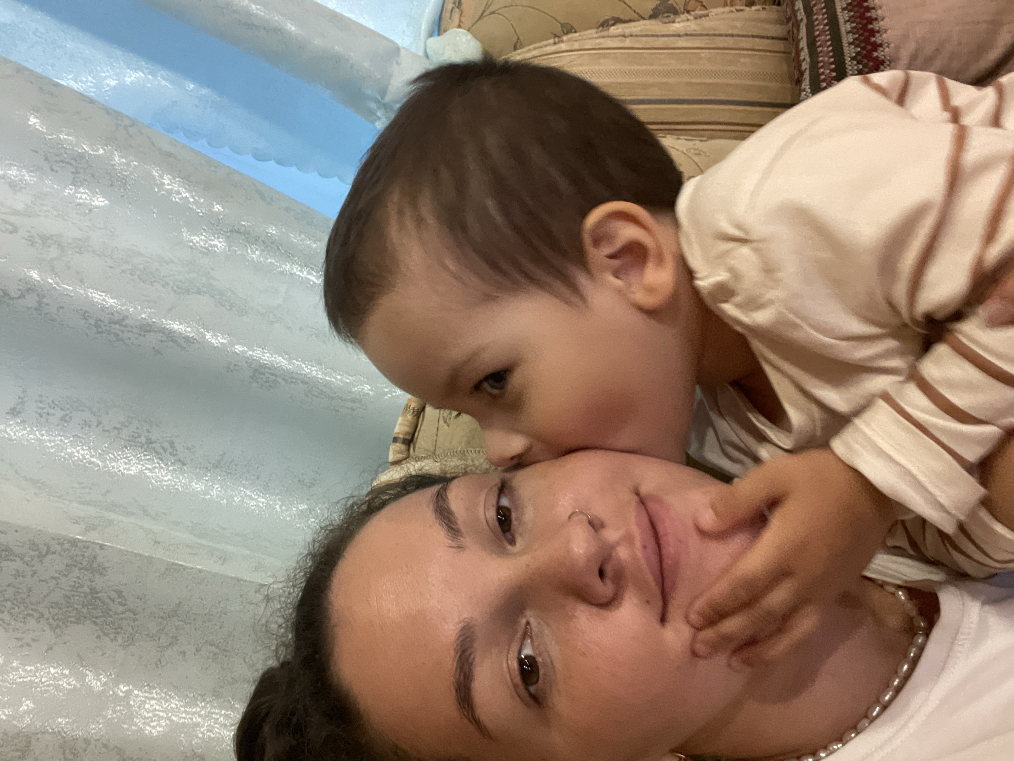 The image shows a close-up, eye-level shot of a woman with her hair pulled back in a bun, wearing a white t-shirt and a pearl necklace. She is looking towards the camera with a slight smile. A young child is kissing her on the cheek, with their hand on her chin. The child is wearing a striped shirt. They are both positioned in front of a patterned couch with a curtain in the background. The lighting is warm and soft.