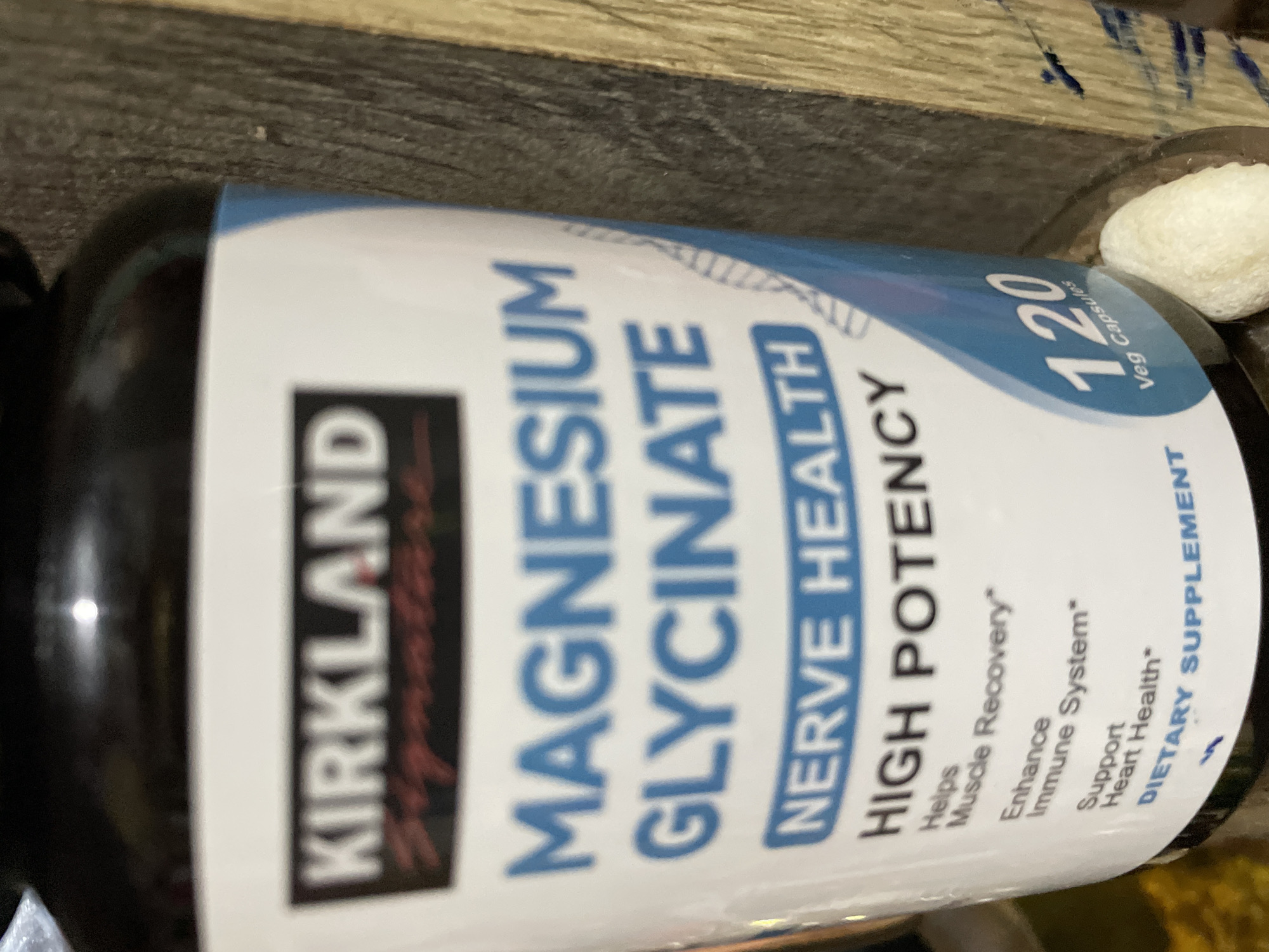 The image displays a close-up shot of a bottle of Kirkland Signature Magnesium Glycinate dietary supplement, positioned on a dark wooden surface in what appears to be an indoor setting in Davao City, Philippines. The bottle is dark-colored with a prominent white and blue label, oriented diagonally across the frame.
The label clearly identifies the product as "KIRKLAND Signature MAGNESIUM GLYCINATE," emphasizing "NERVE HEALTH" and "HIGH POTENCY." Smaller text on the label highlights its benefits: "Helps Muscle Recovery*", "Enhance Immune System*", and "Support Heart Health*." It is further categorized as a "DIETARY SUPPLEMENT." The top right portion of the label indicates that the bottle contains "120 Veg Capsules."
To the right of the bottle, partially visible, is a small, irregular-shaped white object, possibly a piece of cotton or a similar soft material. The background is a dark, wood-grained surface, which looks like a table or counter. The lighting is artificial, causing some glare on the bottle's surface, particularly near the top. No people are visible, and no specific activity is taking place beyond the still-life presentation of the product. The overall composition is a product photograph, possibly for e-commerce or personal inventory.