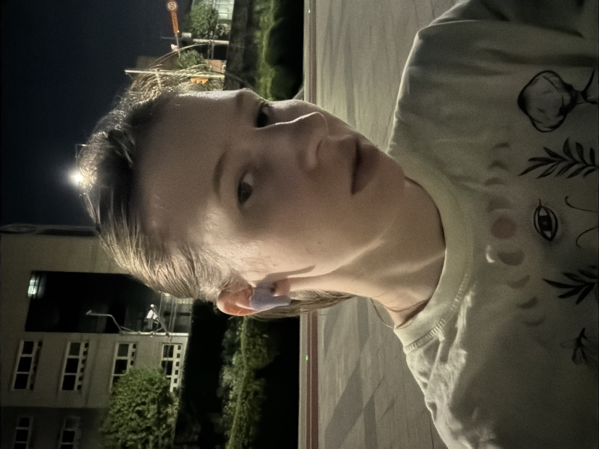 A young person with light-colored hair pulled back and a purple earbud in their right ear is looking towards the camera. They are wearing a light-colored t-shirt with a celestial design. The background shows a multi-story building with windows, lush green bushes, and a street or paved area. The scene appears to be outdoors at night, illuminated by artificial lights. The location is identified as Dongducheon-si, South Korea.