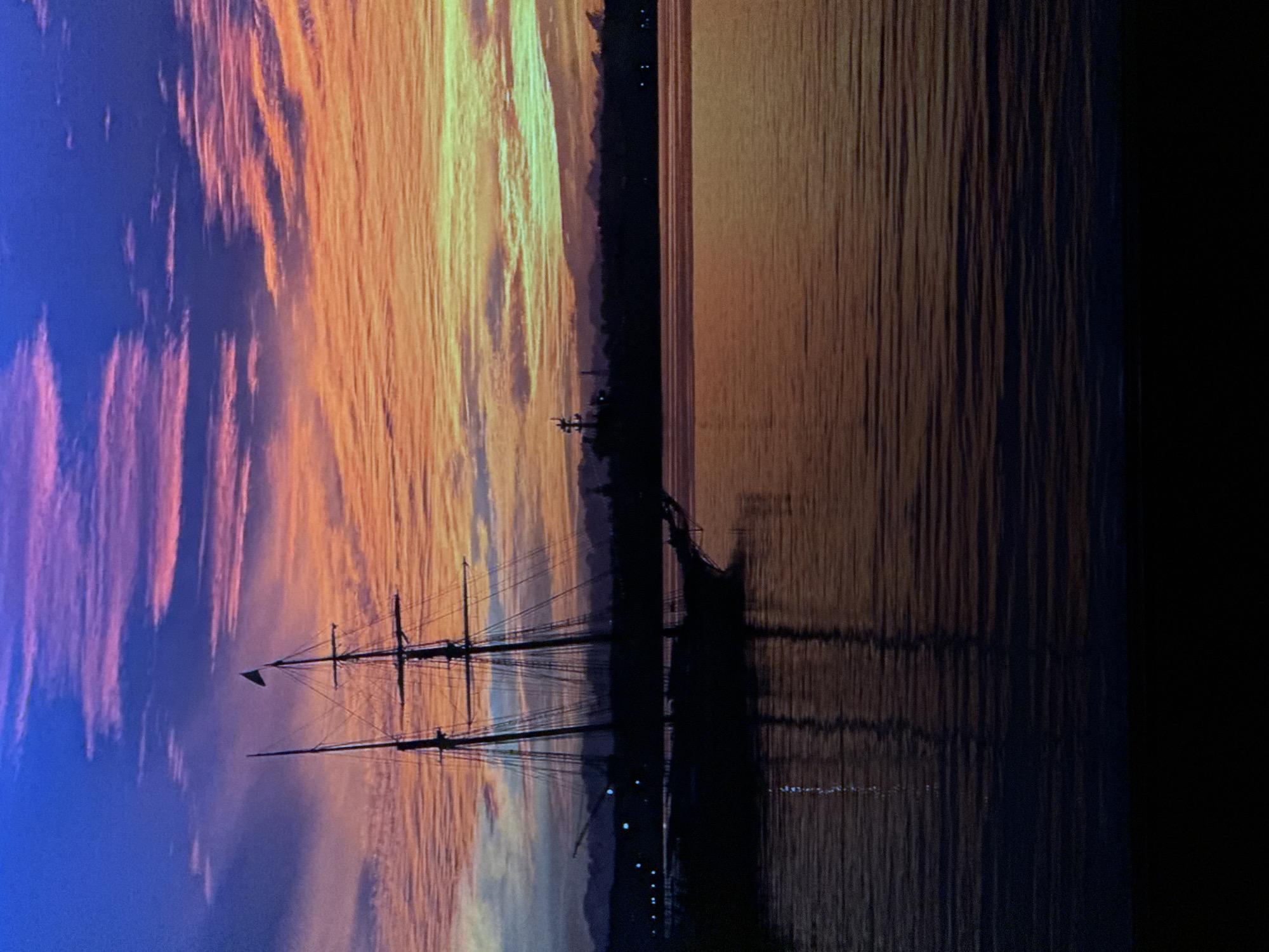 The image depicts a sunset scene over a body of water, with a silhouetted sailing ship in the foreground. The sky is a vibrant display of colors, transitioning from deep blue at the top to shades of purple, pink, orange, and yellow towards the horizon. Wispy clouds are visible, catching the light of the setting sun. The water reflects the colors of the sky, creating a shimmering surface. In the distance, a dark silhouette of land, possibly a city skyline with a few lights, is visible. There are no people or animals visible in the image. The overall mood is serene and picturesque. There is no information or visual cues to suggest the location is Saratov, Russia.