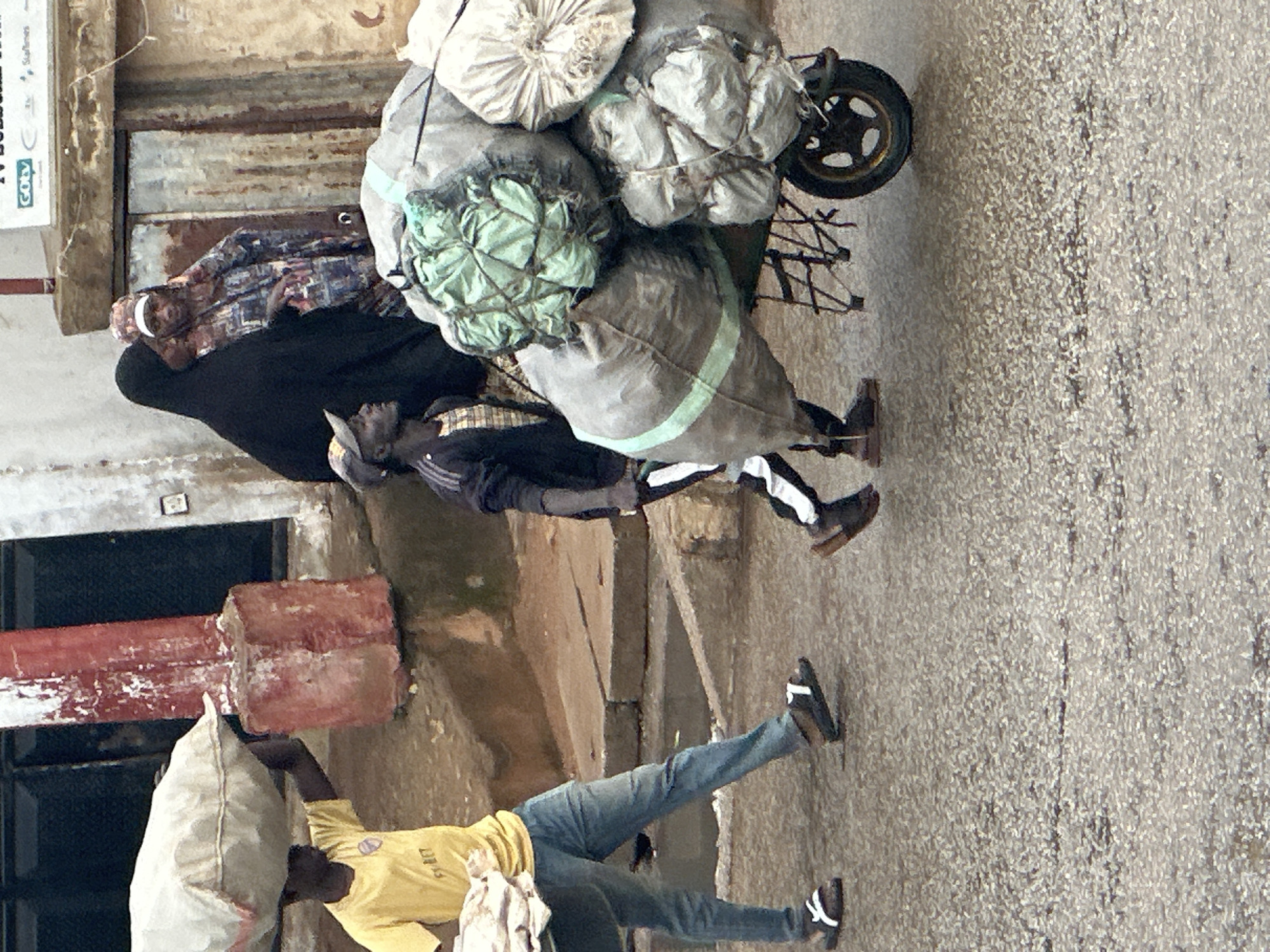 The image shows a street scene in Bukuru, Nigeria, with several people engaged in transporting goods. In the foreground, a person wearing a yellow t-shirt and blue jeans carries a large white sack over their shoulder. Behind them, another person is walking, also carrying a sack on their head. Further back, a person in a black garment and a checkered shirt is pushing a cart overloaded with large, bundled items, some wrapped in green netting. The cart has a single wheel and a metal frame. The setting appears to be a dusty, outdoor environment with buildings and walls visible. The lighting suggests it is daytime, possibly overcast or late afternoon given the muted tones. The overall impression is one of hard work and labor, with individuals focused on moving their cargo through the urban landscape. There is a sign visible on a white surface in the upper left portion of the image, but the text is not clearly legible.