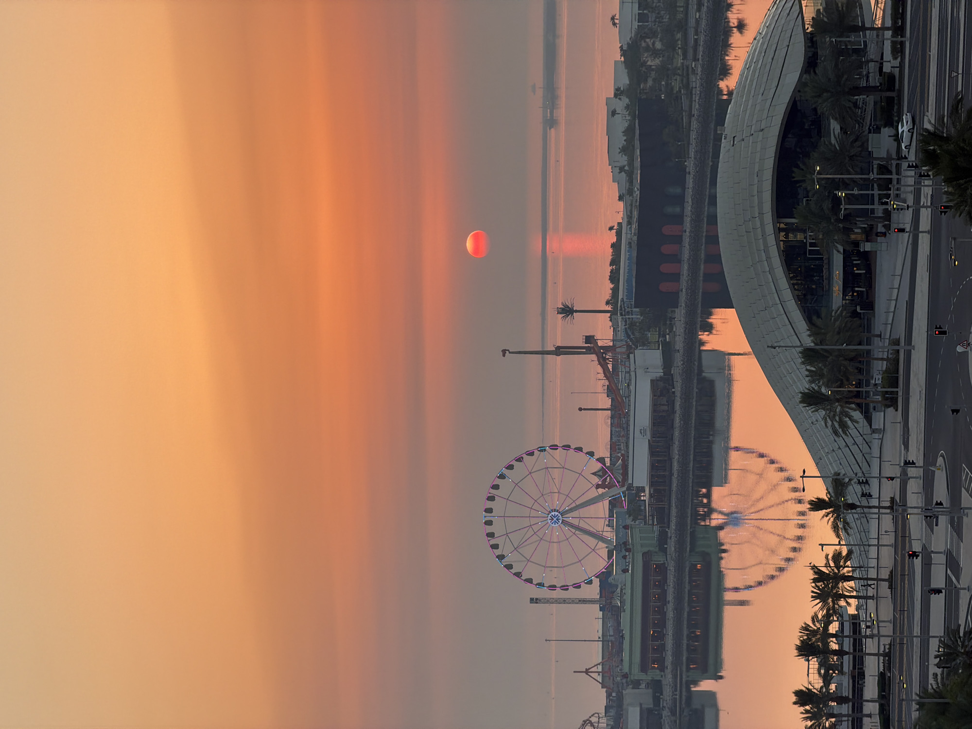 An aerial, medium shot captures a vibrant sunset over a waterfront amusement park and modern architectural structures. The sky is a gradient of warm oranges and pinks, with the sun a bright red disc hovering near the horizon. The water reflects the colorful sky and the lights of a large Ferris wheel, which is illuminated with pink and blue lights. Several other amusement park rides are visible, including roller coasters and tall towers. In the foreground, a contemporary building with a curved roof is surrounded by palm trees. The scene is set in an urban coastal area, with a body of water separating the amusement park from the foreground buildings. The overall impression is one of leisure and urban development against a dramatic natural backdrop.