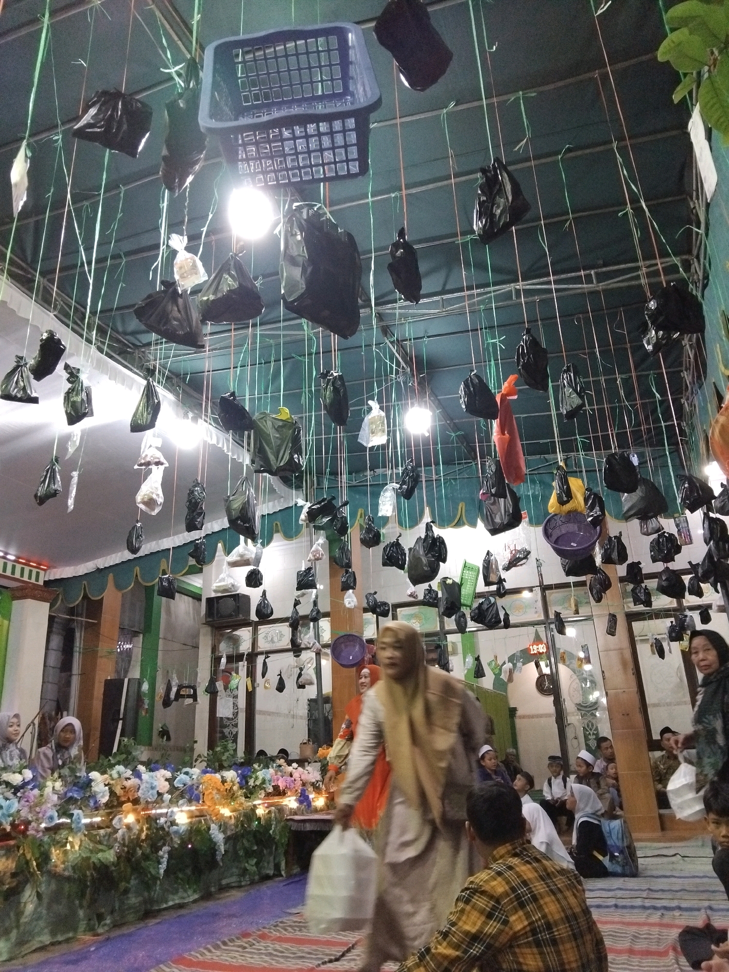 In a dimly lit indoor space, possibly a community hall or mosque in Kepuh Kiriman, Indonesia, a unique arrangement of objects hangs from the ceiling by strings. These objects consist of numerous black plastic bags, some appearing to contain items, suspended at various heights. A few clear plastic bags are also visible, along with a blue plastic basket and a purple plastic bowl. Green and light green strings create a dense network above. The ceiling itself appears to be made of corrugated metal, with exposed beams.
Below this overhead display, the scene is populated by several individuals. In the foreground, a person in a yellow and black checkered shirt is seen from behind, facing away from the camera. To their left, a person in a light brown hijab and robe is walking away from the camera, carrying a white plastic bag. Further left, two young girls in hijabs are standing near a decorative arrangement of artificial flowers and lights. To the right, a woman in dark clothing is walking towards the camera, also carrying a white bag. Other individuals, including children and adults, are visible in the background, some seated and others standing, with some wearing traditional Islamic headwear. The floor is covered with a striped carpet. The overall atmosphere suggests an event or gathering is taking place.