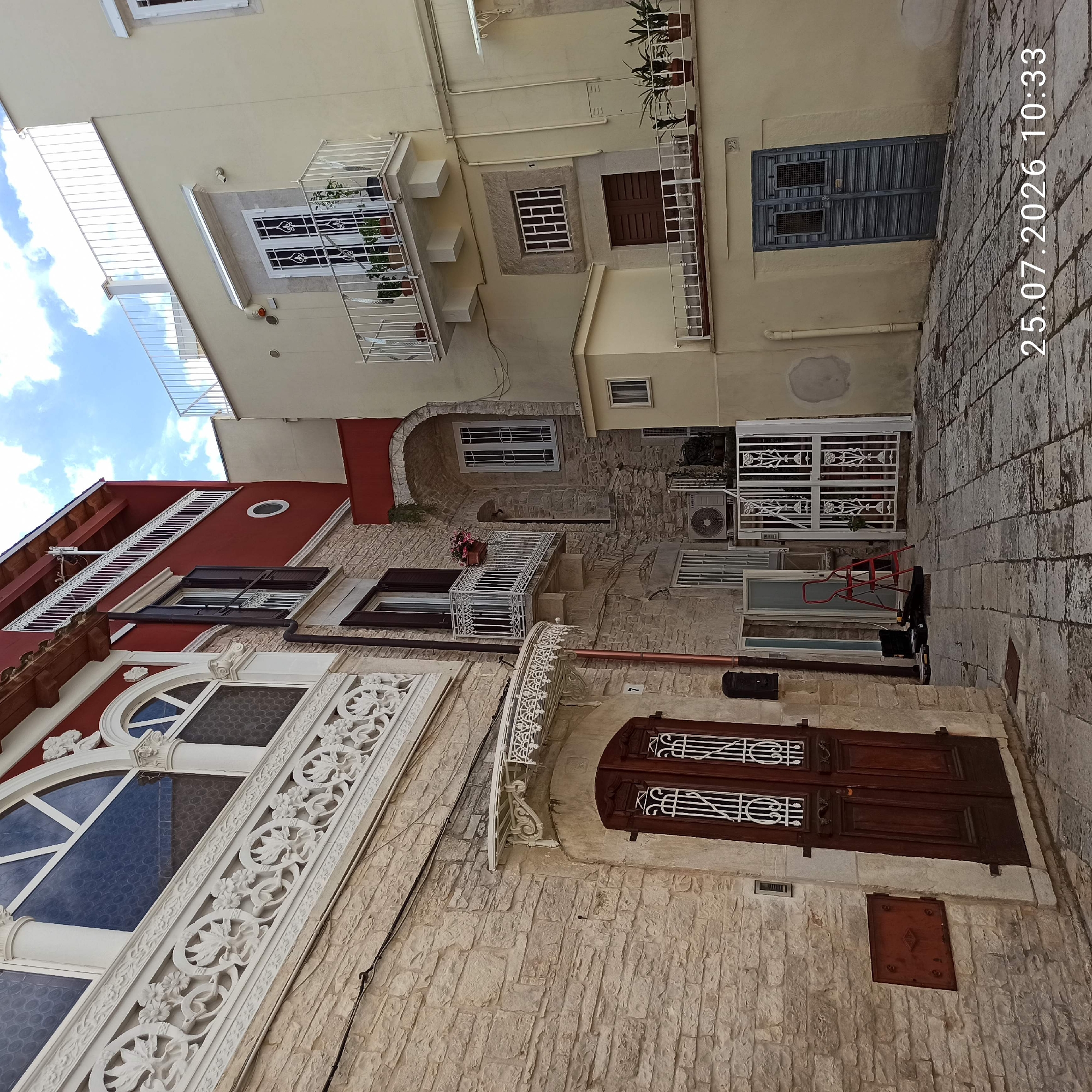 The image shows a narrow, cobblestone street in Ruvo di Puglia, Italy. On the left is a light-colored stone building with a large, dark wooden double door featuring ornate white metal detailing. Above the door, an ornate white metal frieze runs horizontally across the building. Adjacent to this, a red-colored building with a white balcony and dark shutters is visible. On the right is a cream-colored building with a white balcony adorned with potted plants. Further down the street, a dark blue double door is set into the cream-colored building. The scene is documented with a timestamp of 25.07.2026 10:33. The sky above is partly cloudy.