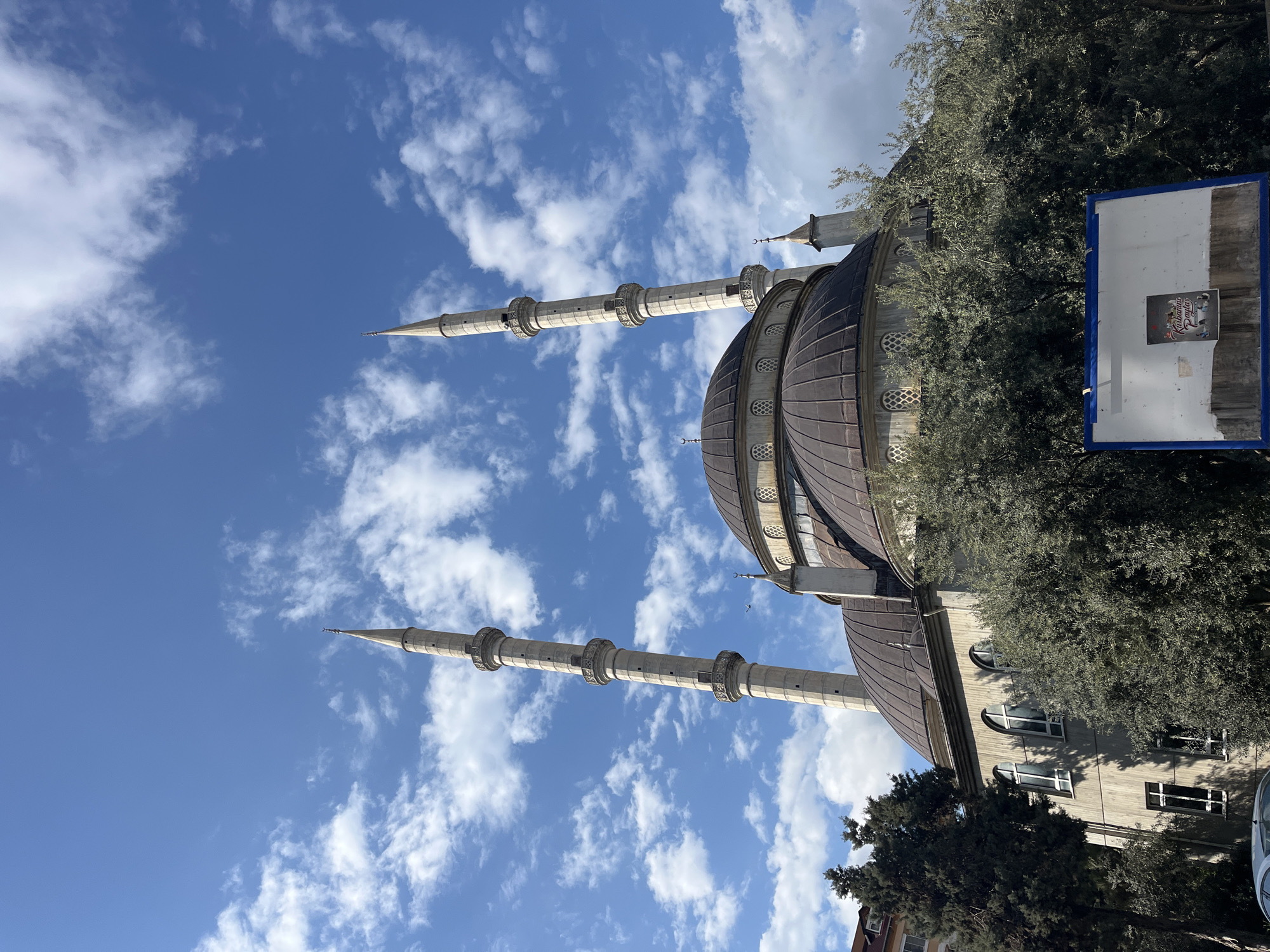 Here is a description of the image:
The image shows a mosque with two minarets against a partly cloudy blue sky, possibly in Alanya, Turkey. The mosque features a large, dark-colored dome and tall, cylindrical minarets with decorative elements. Part of the building is obscured by trees. A basketball backboard and a building are located behind the mosque, adding to the context. The image is rotated 90 degrees clockwise, and the perspective is from a low angle, emphasizing the height of the mosque and minarets.