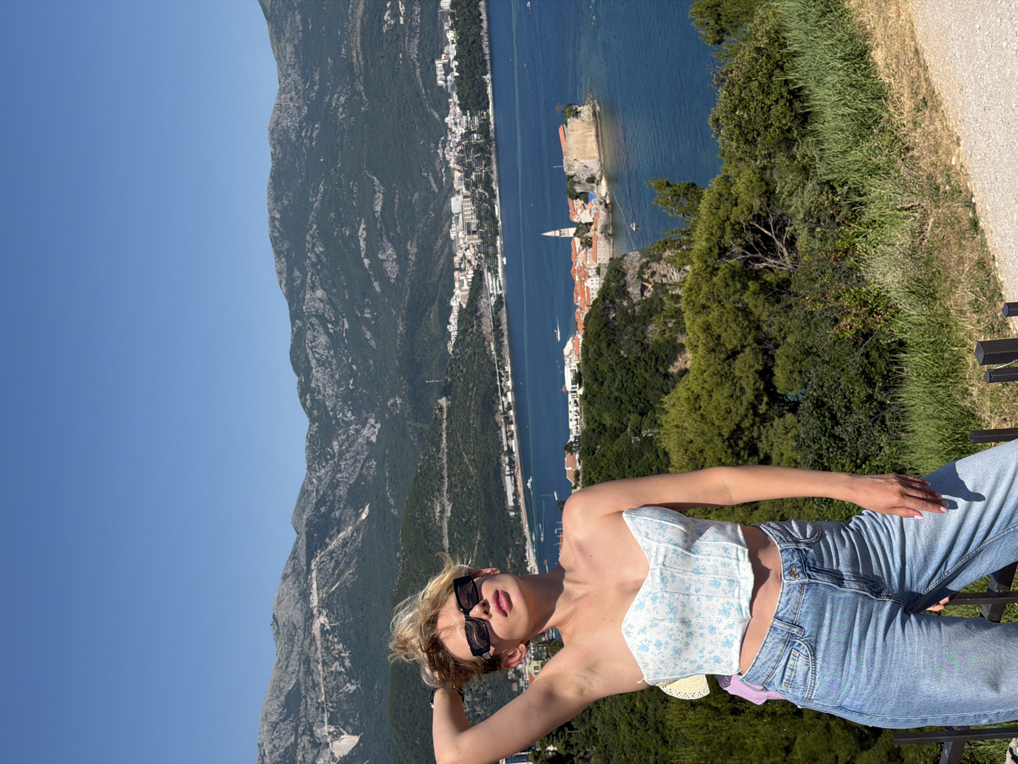 A young woman stands in the foreground, positioned slightly left of center, against a panoramic view of Budva, Montenegro. She has blonde hair styled loosely, wears black rectangular sunglasses, a white strapless corset top with light blue floral patterns, and light blue denim jeans. Her left arm is raised with her hand behind her head, while her right arm is at her side. Her head is tilted slightly upward and to the right, revealing pink lipstick.
The background features a clear blue sky occupying the top quarter of the image, followed by a range of grey and green mountains. Below the mountains, the city of Budva is visible along the coastline, with numerous buildings extending down to the water. A large expanse of deep blue water, identified as the Adriatic Sea, stretches across the middle ground, containing several small boats. A prominent fortified peninsula or island, consistent with Budva Old Town (Stari Grad), is situated in the mid-right, featuring red-roofed buildings and a distinct church tower. The immediate foreground consists of dense green foliage, including trees, bushes, and grass, sloping down toward the sea. A light brown dirt path or road is visible in the bottom right corner, alongside a dark fence-like structure in the lower left.
