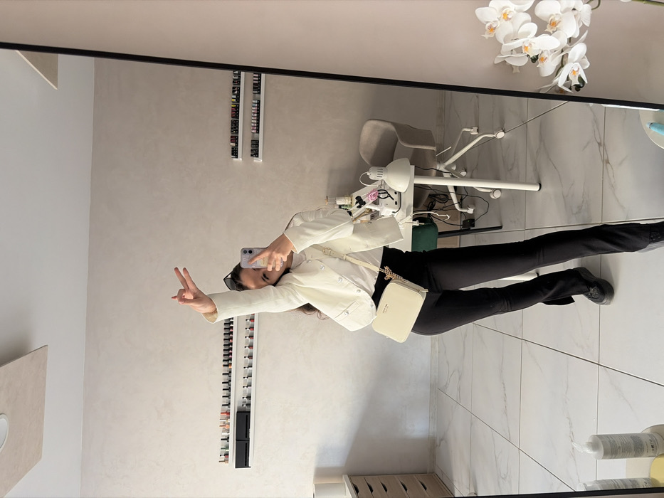 A female individual is captured in a full-length mirror reflection within an indoor setting identified as a nail salon or beauty studio in Sofia, Bulgaria. She is actively engaged in taking a selfie using a smartphone held in her left hand, which partially obscures her face. Her right arm is raised, with fingers forming a "peace" sign. She is dressed in a white short jacket over a white top, black fitted pants with subtle split hems, and black platform shoes. A cream-colored cross-body bag with gold-colored chain accents is slung over her left shoulder. Dark sunglasses rest atop her head.
The background, visible in the reflection, features light beige textured walls and white marble-patterned floor tiles. Two white floating shelves are mounted on the left wall, displaying multiple rows of small bottles, consistent with nail polish or gel. To the right, a white desk with a white five-star wheeled swivel chair is visible. On the desk, a white desk lamp with a conical shade is present, alongside various small bottles, containers, and tools indicative of beauty or nail services. A dark green stool is partially visible beneath the desk. A segment of white artificial orchids is reflected along the right edge of the mirror. A rectangular white light fixture is mounted on the ceiling.