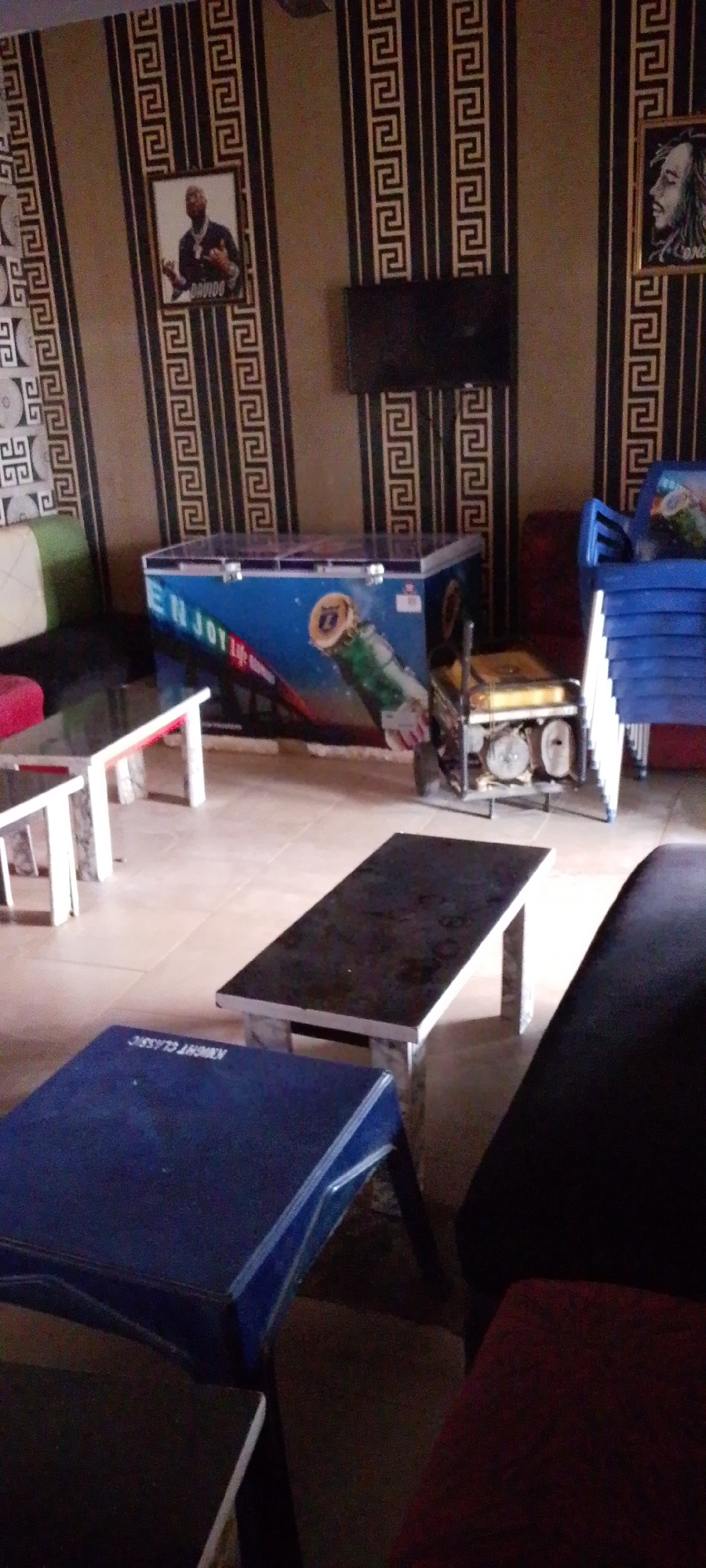The image depicts the interior of an establishment with patterned wallpaper and tiled flooring. In the foreground, there are several tables, including a blue table in the lower left corner with white trim, and a dark rectangular table with a reflective surface supported by stone-like legs. In the middle ground, a large blue cooler with "ENJOY" written on it and an image of a beer bottle is present, next to a yellow generator. Further back, a stack of blue plastic chairs is visible. On the wall, there are two framed pictures, one featuring a man and the other a profile of a person with dreadlocks. A dark, flat-screen television is mounted on the wall between the two framed pictures. The environment appears to be a casual indoor space, possibly a bar or lounge.