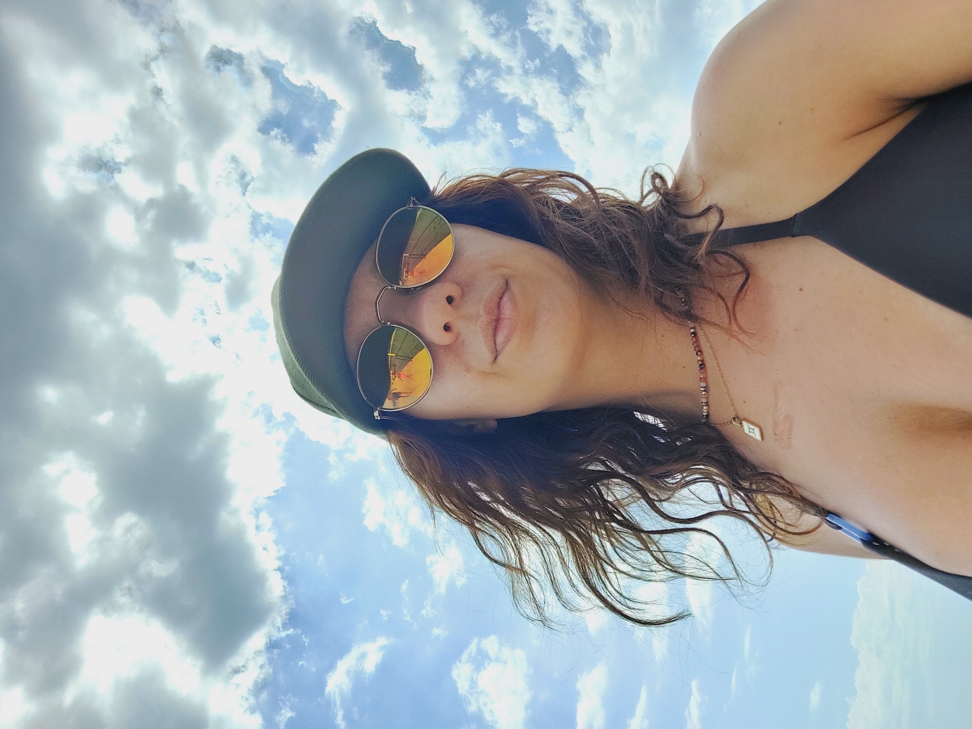 A woman is taking a selfie outdoors against a cloudy sky. She is wearing a dark bikini top, a necklace with a pendant, round sunglasses with reflective lenses showing a reflection of people, and a green baseball cap. Her brown hair is blowing in the wind. The location is Nesebar, Bulgaria.