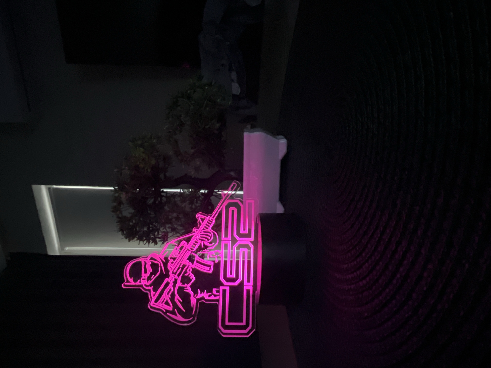This media file shows a neon-style acrylic light display with a soldier figure holding a rifle and the numbers "2024" in the foreground. The figure and numbers are illuminated with a vibrant pink neon light, creating a stark contrast against the dark background. Behind the display, a small artificial tree with green foliage is visible, positioned near a white rectangular object that appears to be part of the light's stand or housing. In the upper background, a dimly lit, dark abstract form can be seen, possibly a sculpture or part of the room's decor. The overall setting is dark, suggesting it is nighttime or an indoor space with minimal lighting. The focus is on the glowing display, which has a modern and somewhat edgy aesthetic. The image is taken from a low angle, with the display filling the lower portion of the frame. The lighting creates a dramatic effect, highlighting the outline of the soldier and the bold numerals. There is no discernible weather information or specific location cues beyond the provided context of Varna, Bulgaria. The mood conveyed is one of futuristic or stylized representation, with the soldier motif potentially alluding to themes of protection, year-end commemoration, or gaming culture.