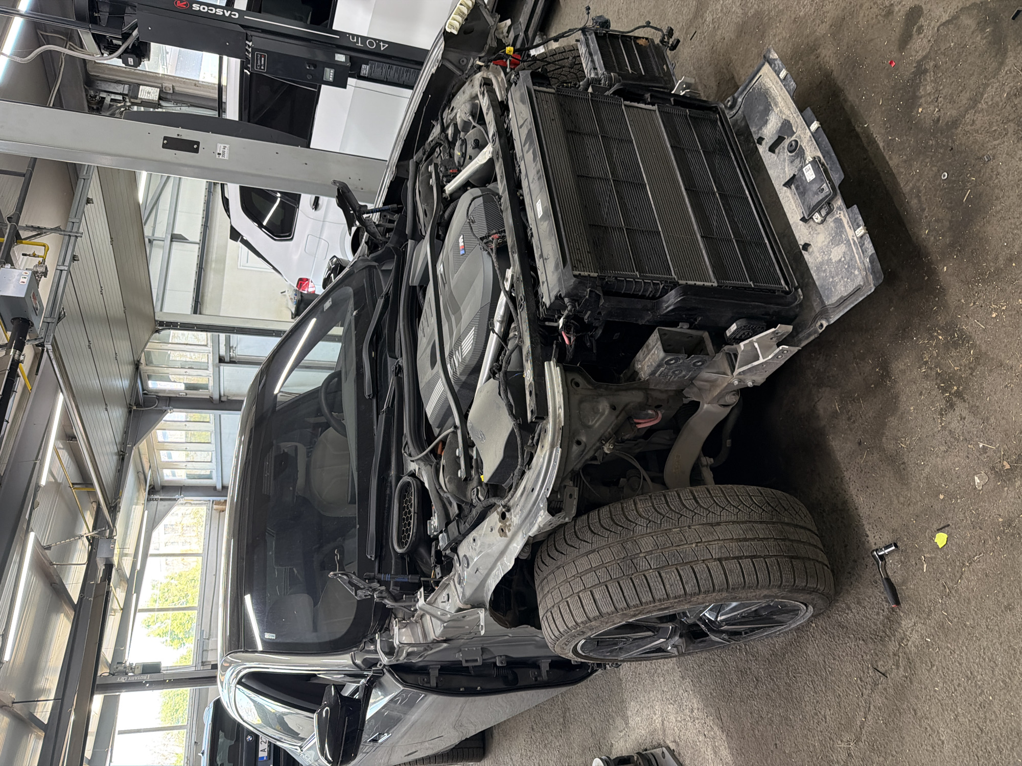 The image shows a partially disassembled car, likely a BMW, in a garage setting. The front end of the car is removed, exposing the engine and radiator. A tire is visible on the right side of the frame. The garage has a lift, and other equipment is present. The location is unknown and there is no evidence of being in Burgas, Bulgaria.