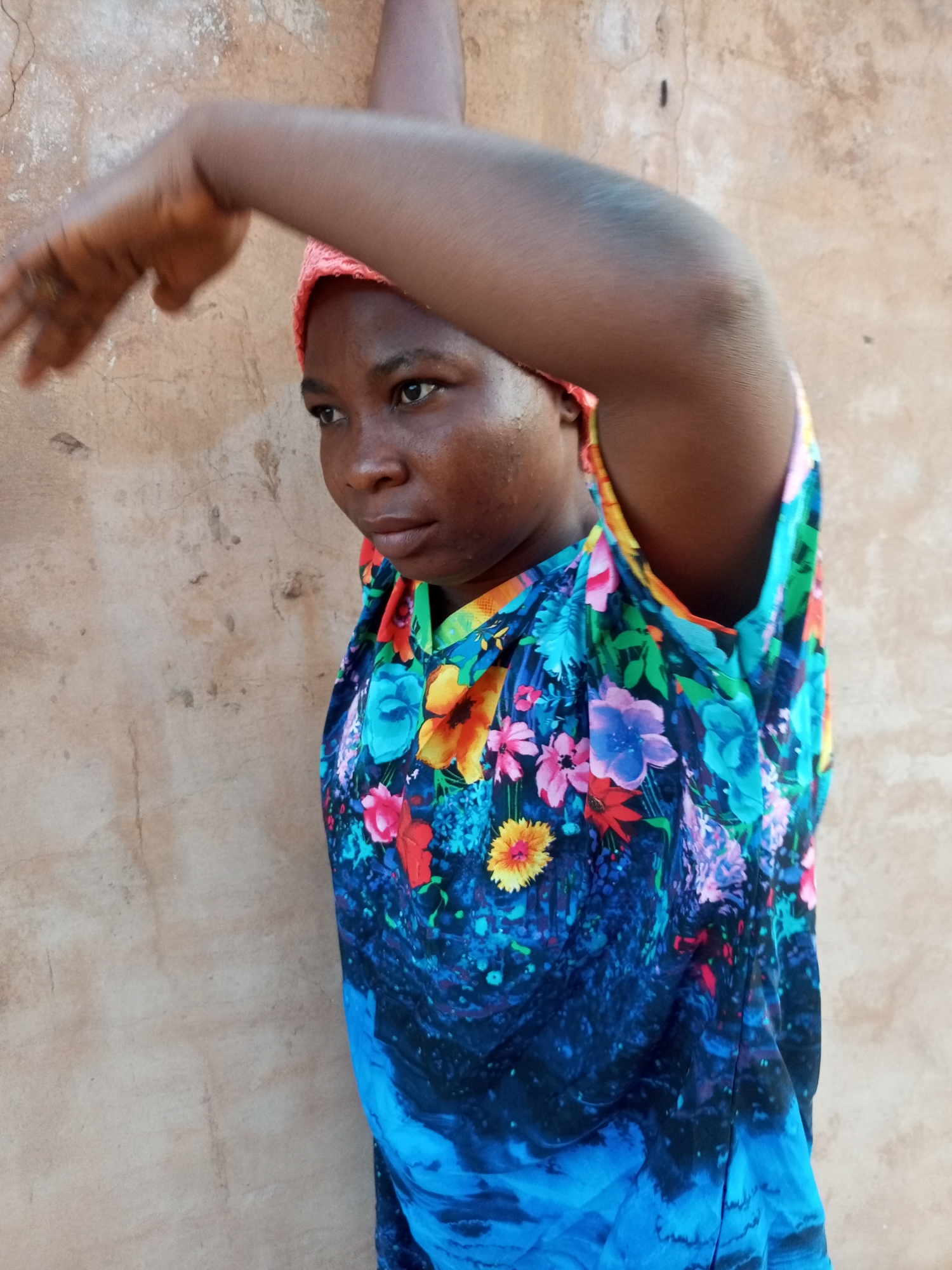A young Black woman stands against a weathered, light brown wall. She is wearing a vibrant, floral-patterned dress with shades of blue, green, yellow, and pink, and a pink headscarf. Her right arm is raised, with her hand extended towards the left, partially obscuring her face. Her head is turned to her left, and she appears to be looking downwards and to the left. Her skin has a slight sheen, suggesting sweat or oil. The background wall has a rough, textured surface with some dark marks and cracks. The lighting suggests it is daytime, with some shadows cast on the wall.