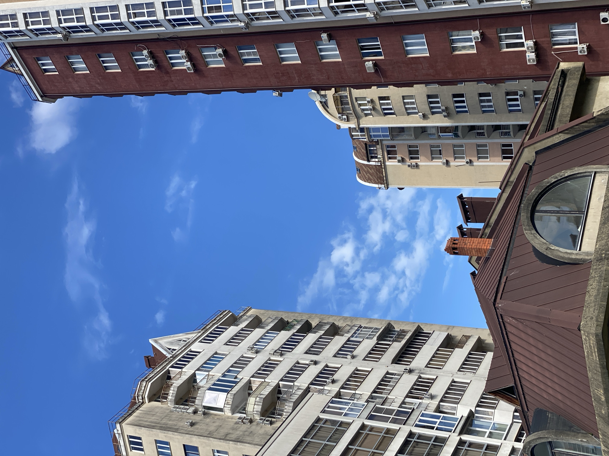 The image captures a view of several buildings in the city of Sochi, Russia, looking upwards towards the sky. The sky is a clear blue with some wispy white clouds. The buildings are of varying heights and architectural styles.
The top of the image features a reddish-brown building with rows of windows and air conditioning units. Below that is a building with a light beige color scheme, also with windows and air conditioning units.
In the lower part of the image, a white building is visible with numerous windows and projecting balconies. To the right is a brown building with a distinctive round window and a visible chimney.
The image is taken from a low angle, emphasizing the height of the buildings. The overall composition creates a sense of urban density and architectural diversity.