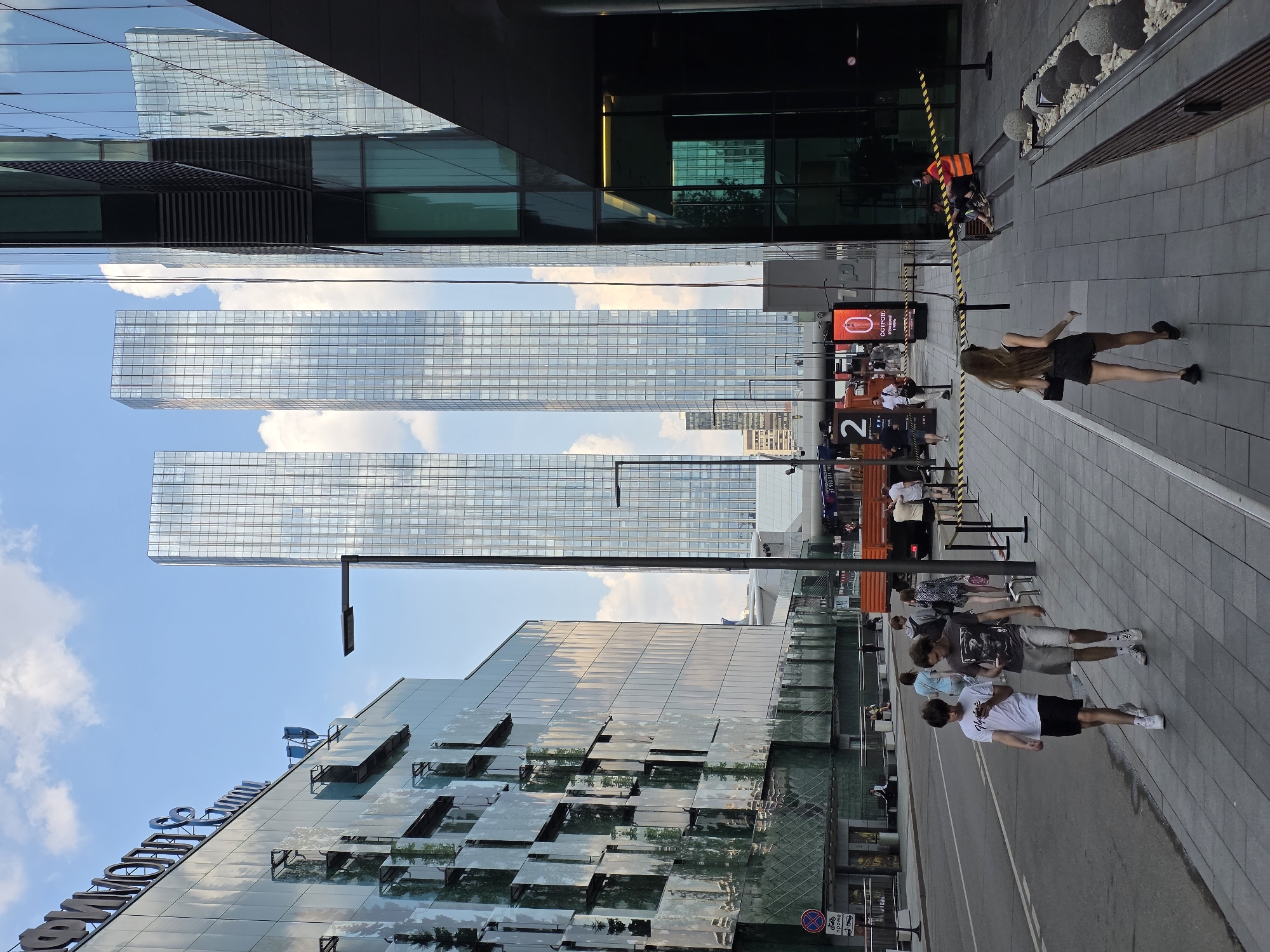 The image shows a street scene in Moscow, Russia, likely in a modern business district given the presence of glass skyscrapers and contemporary architecture. In the foreground, two young males are walking on a sidewalk, each engrossed in their mobile phones. The male on the left wears a white t-shirt and black shorts, while the one on the right wears a graphic t-shirt and grey shorts. Behind them, a young female with long brown hair, wearing a black tank top and shorts, walks away from the camera.
Further down the sidewalk, several other individuals are visible. A person in an orange safety vest sits on a low wall. To the left, a street with a road and a sidewalk stretches into the distance, flanked by large, modern glass buildings. A red truck with the number "2" displayed on its side is parked on the street. Two tall, modern glass skyscrapers with a grid-like facade dominate the background, reflecting the blue sky and clouds. The environment appears to be an urban setting with clear weather.