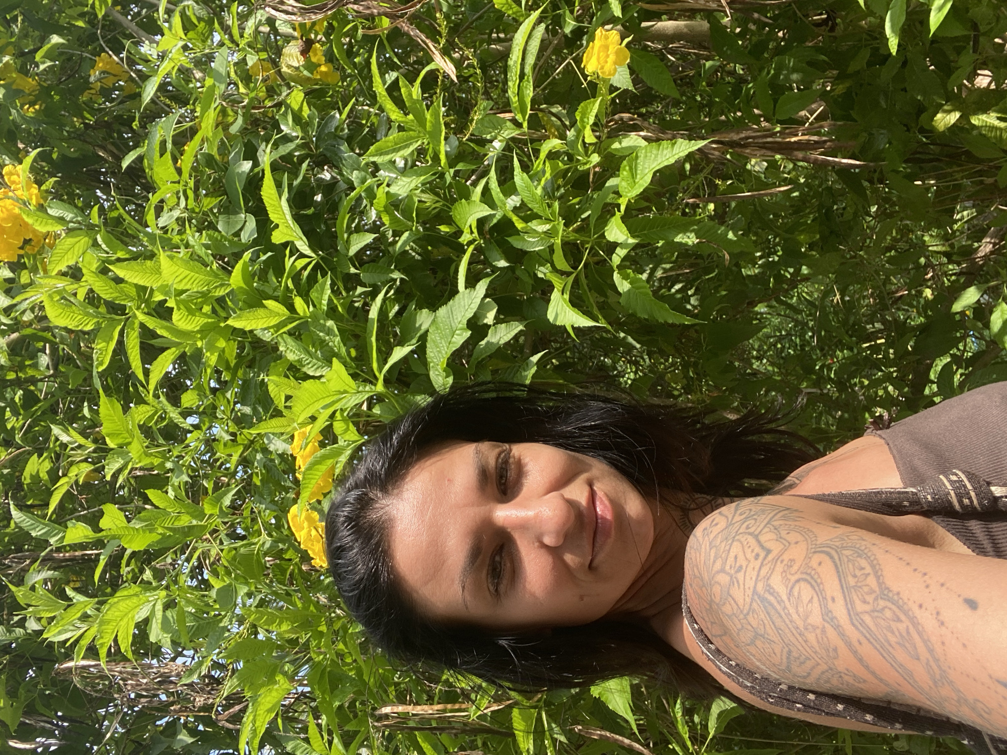 A woman with dark hair and a tattoo on her left arm takes a selfie in front of a dense, green bush with yellow flowers. She is wearing a brown top. The background consists of foliage and branches. The image appears to be taken outdoors, possibly in a garden or natural setting. The location is Mũi Né, Vietnam.