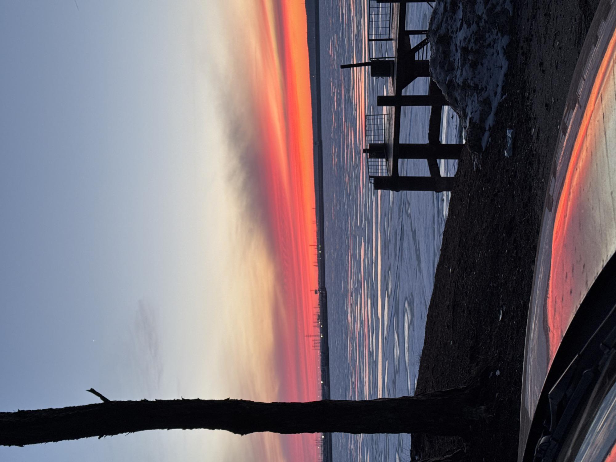 The image shows a sunset over the Kama River in Chaikovsky, Russia. The sky is a gradient of colors from blue to orange to red. There is an industrial area on the horizon with power lines and buildings. The river reflects the colors of the sunset. On the right side of the image is the edge of a vehicle (possibly a car) and some snow-covered ground. A metal pier or structure is visible along the riverbank. A silhouetted tree branch is in the lower part of the image.