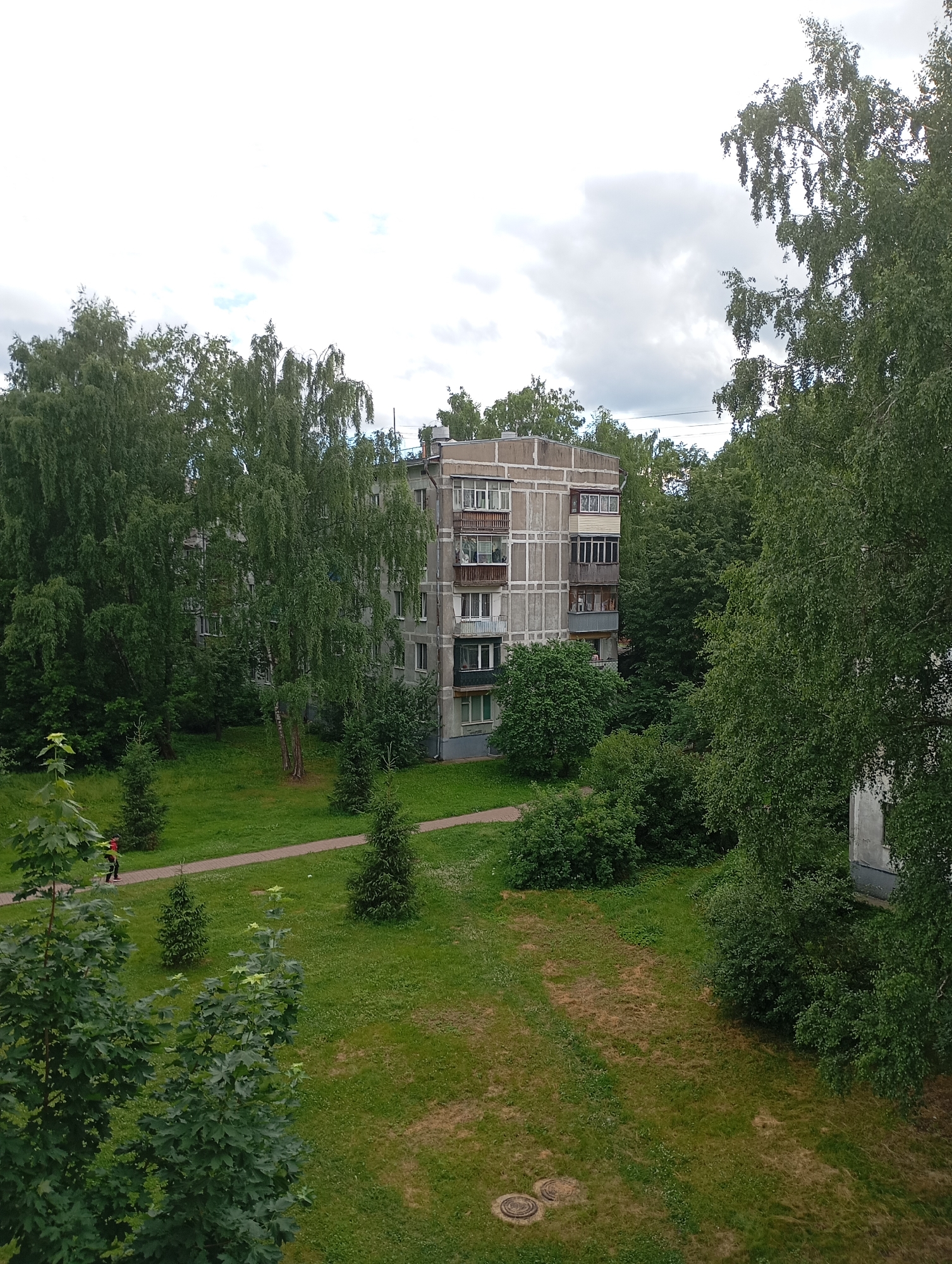 This is an outdoor, eye-level shot of a residential building in Gorki-2, Russia. The building, constructed of concrete panels, features multiple floors with balconies and windows. It is surrounded by lush green trees and grass. A paved walkway runs through the grassy area. A person wearing an orange jacket and dark pants is walking on the path. The sky is overcast with clouds.