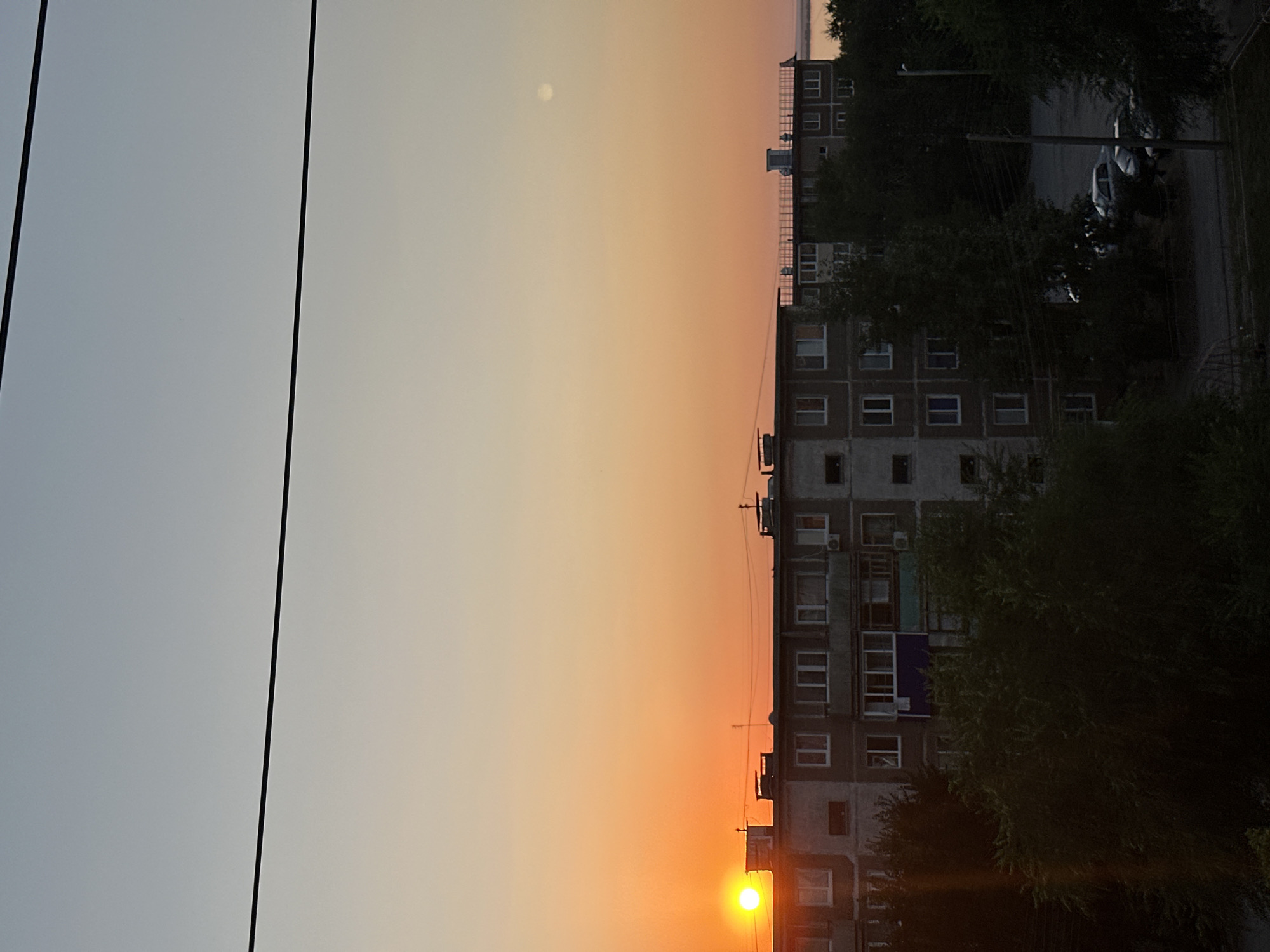 A sunset over the city of Energetik, Russia, casting a warm orange glow across the sky. The sun, a bright orb, is partially obscured by a building on the left side of the frame. In the midground, a multi-story apartment building with many windows stretches across the scene. Trees in the foreground obscure the lower portion of the building and the street below, where several parked cars are visible. The sky is a gradient of soft colors, transitioning from a pale blue at the top to a vibrant orange near the horizon. A thin, hazy moon or planet is visible in the upper right corner of the sky. Electrical wires are strung across the top of the image, adding a man-made element to the natural beauty of the sunset.
