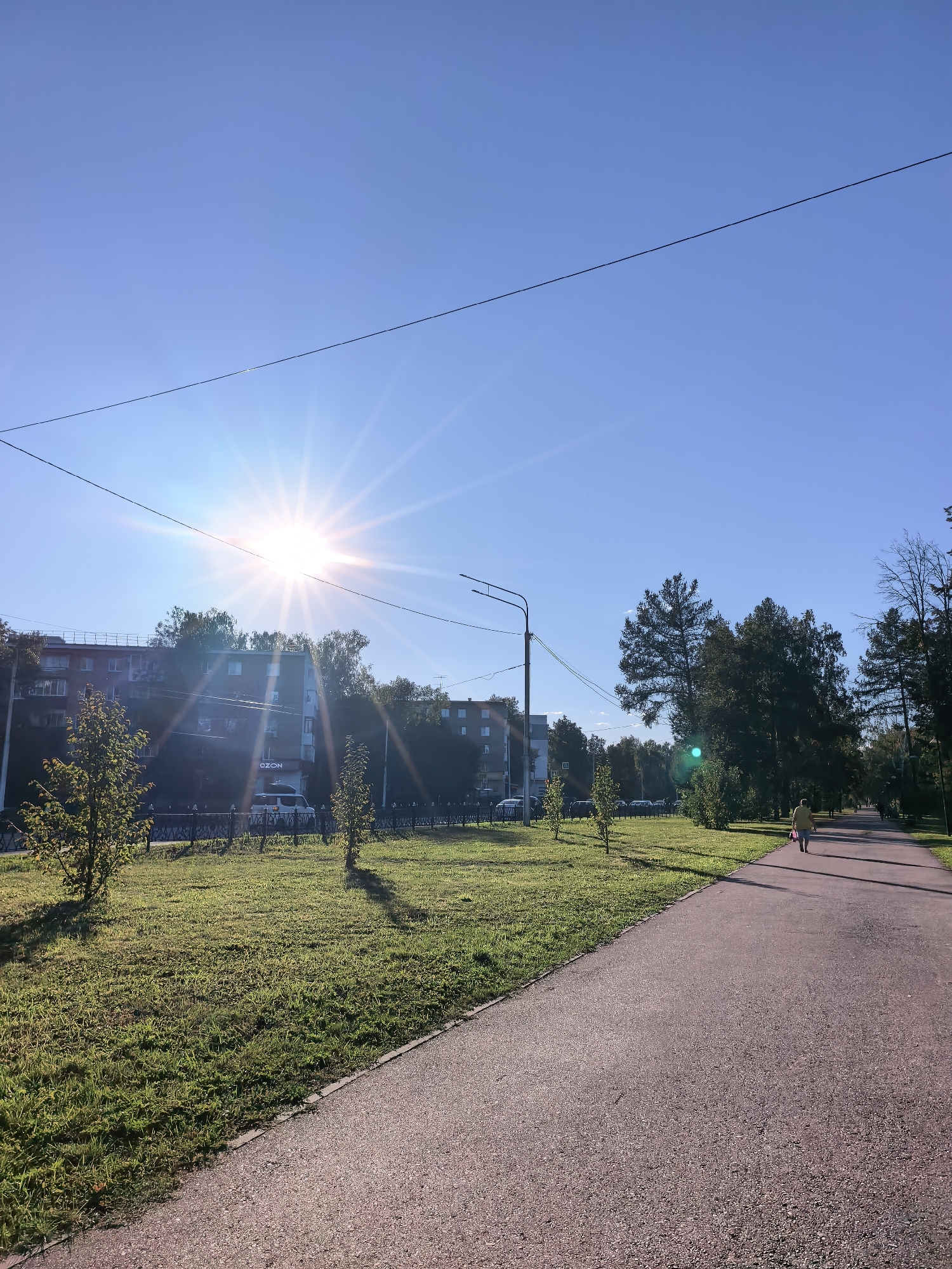 This is a vertical, eye-level, full shot of a sunlit park path in Sterlitamak, Russia. The sun is high in the sky and to the left, casting bright rays across the scene. A paved path runs from the foreground to the middle ground on the right. On the left side of the path is a grassy area with scattered trees and a low black fence. Behind the fence, there is a multi-story apartment building with windows. Beyond the apartment building, there are more trees. A single person, wearing a yellow shirt and carrying a pink bag, walks away from the camera on the path in the middle ground. The sky is clear and blue.