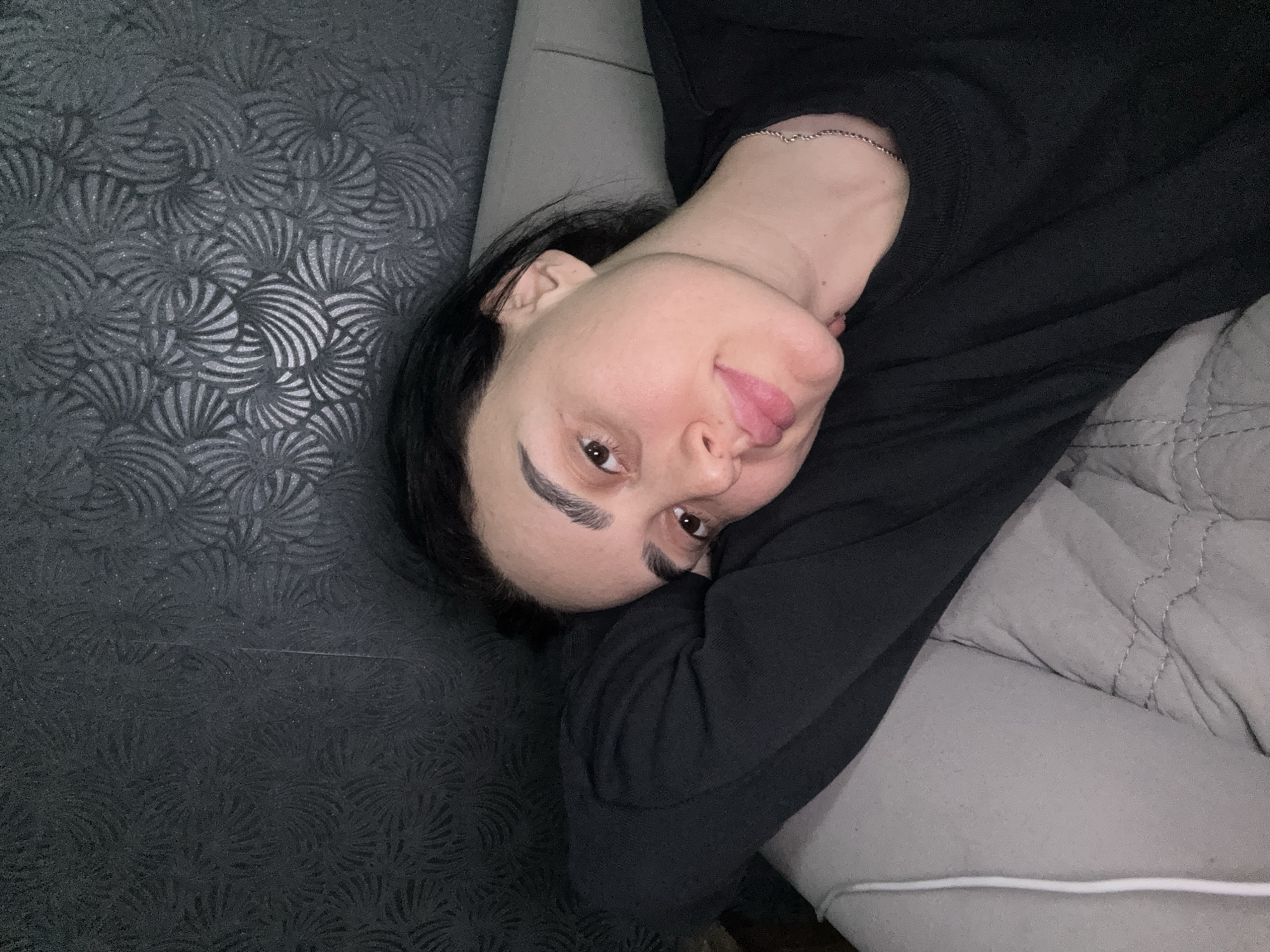 The image displays a person, likely a woman, reclining in a casual indoor setting. She has dark hair, prominent dark eyebrows, and brown eyes, looking directly at the camera with a gentle, relaxed smile. A thin silver chain is visible around her neck, and she is wearing a plain black long-sleeved top. Her head is tilted, suggesting she is lying on her side.
On the left side of the frame, behind her, is a dark wall or headboard featuring a textured, patterned design. This pattern consists of repeating, fan-like or shell-like motifs, which seem to have a subtle shimmer or glitter that catches the light in places. To her right, a light gray, soft, and possibly quilted surface, resembling a bedspread or sofa cushion, indicates she is resting comfortably.
The lighting appears to be ambient indoor light, creating a relaxed atmosphere. There is no discernible text within the image. The person's expression conveys a sense of ease and contentment. The location context for this scene is Tbilisi, Georgia.