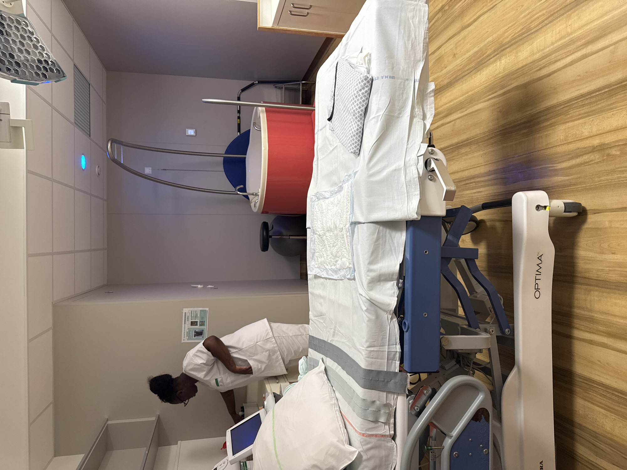 A nurse in a white uniform is standing by a medical bed in a room that appears to be a hospital or birthing center. The room has a clean, sterile environment with medical equipment visible, including a birthing tub with a red exterior and blue interior, a medical bed with white linens, and what looks like a monitor or control panel. The floor is made of light-colored wood. The nurse is looking down at the monitor. The overall scene suggests a medical facility, possibly in Tourcoing, France, given the context of the prompt.