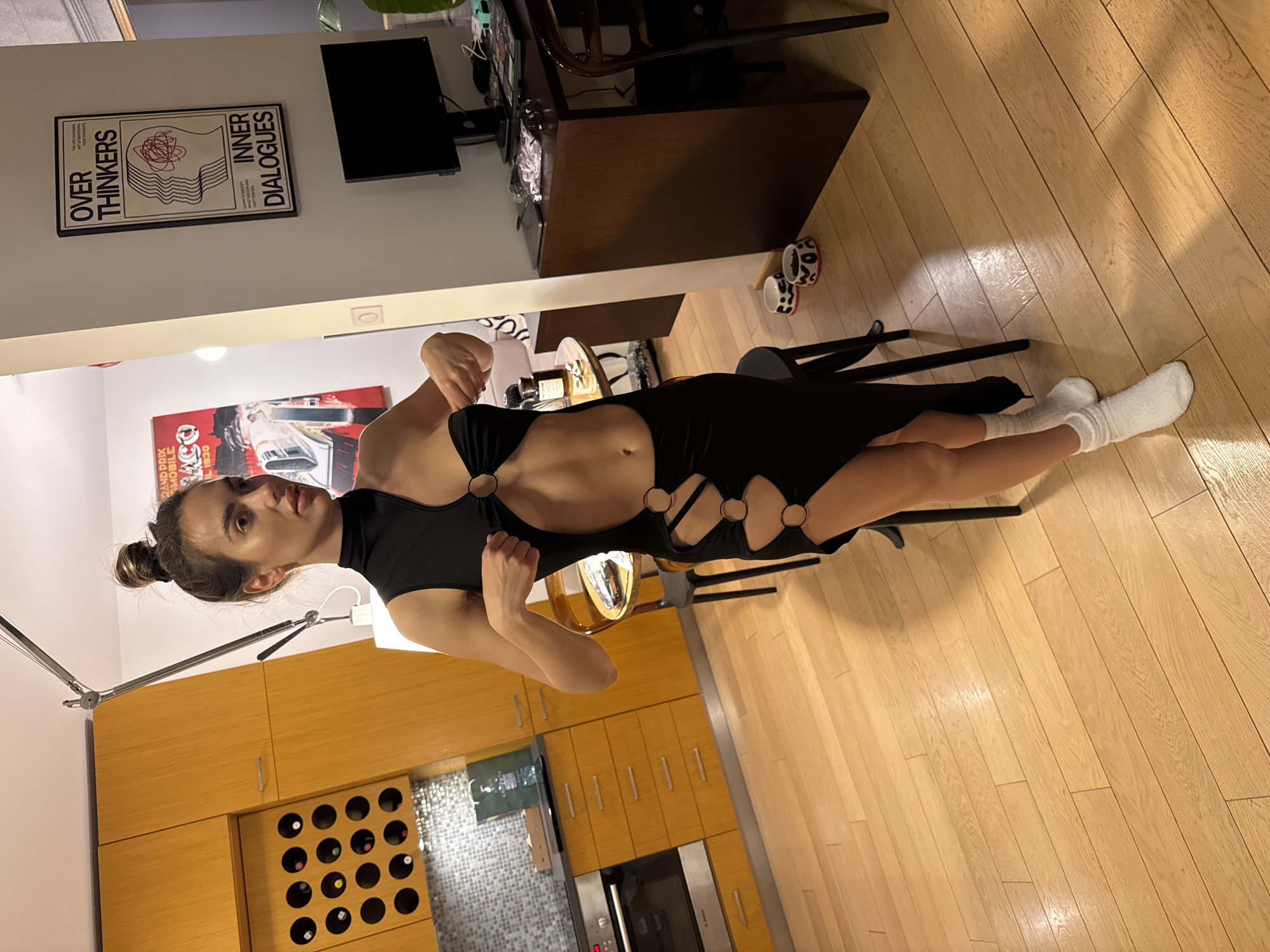 A young woman is standing indoors, likely in an apartment in Warsaw, Poland, given the presence of Polish text in a poster ("GRAND PRIX MOBILE ACO 1930"). She is wearing a black dress with circular cutouts along the sides and white socks. She appears to be posing, with her hands positioned in front of her. The background includes wooden floors, a kitchen area with cabinets and a wine rack, a dining area with a table and chairs, and a living area with a TV and posters. One poster reads "OVER THINKERS INNER DIALOGUES". There are also decorative bowls on the floor near a desk and chair. Lighting fixtures are visible, one of which is casting light on the woman.