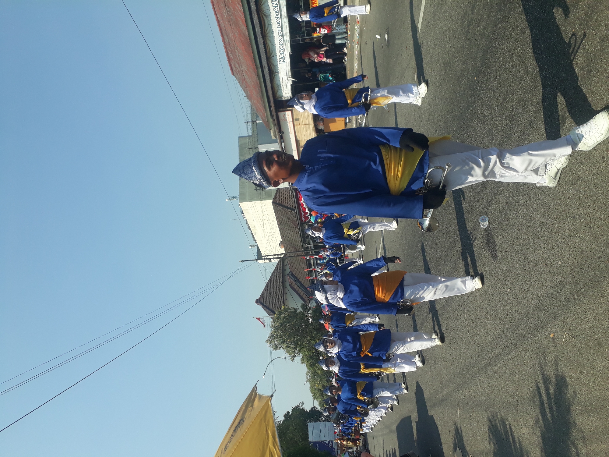 A marching band, composed of individuals wearing blue jackets, white pants, and distinctive blue headwear, proceeds down a paved road under a clear blue sky. The majority of the participants are young men, with some wearing yellow sashes draped over their jackets. Several individuals are holding brass instruments, specifically trumpets, and appear to be marching in formation. In the background, buildings with red roofs and white facades line the street. People are visible on the sidewalks, observing the parade. The scene suggests a public event or festival taking place in a city. The presence of overhead power lines indicates an urban environment. There are some scattered pieces of trash on the road, consistent with a public gathering. The overall impression is one of a daytime outdoor event in a populated area of Pangkalpinang, Indonesia.