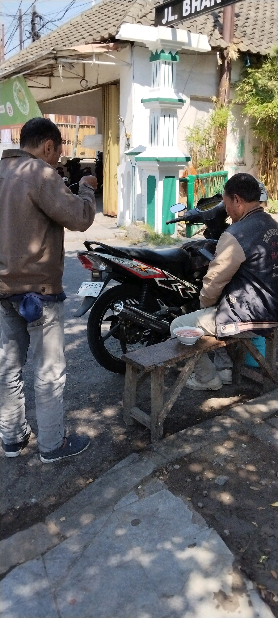 The image depicts an outdoor scene in Surabaya, Indonesia, likely on a street or sidewalk. Two men are present. The man on the left is wearing a brown jacket and grey pants, holding a phone and seemingly interacting with it. He has a blue cap tucked into his back waistband. A motorcycle is parked in front of him. The man on the right is seated on a low wooden stool, wearing a dark jacket with light-colored sleeves and light-colored pants. He is looking down at his phone and has a bowl of food in front of him on the stool. The background shows a building with a tiled roof, a green and white decorative pillar, and some bamboo plants. A street sign with "JL. BH..." is visible above the building. The ground is a mixture of asphalt and concrete. The scene is well-lit, suggesting it is daytime.
