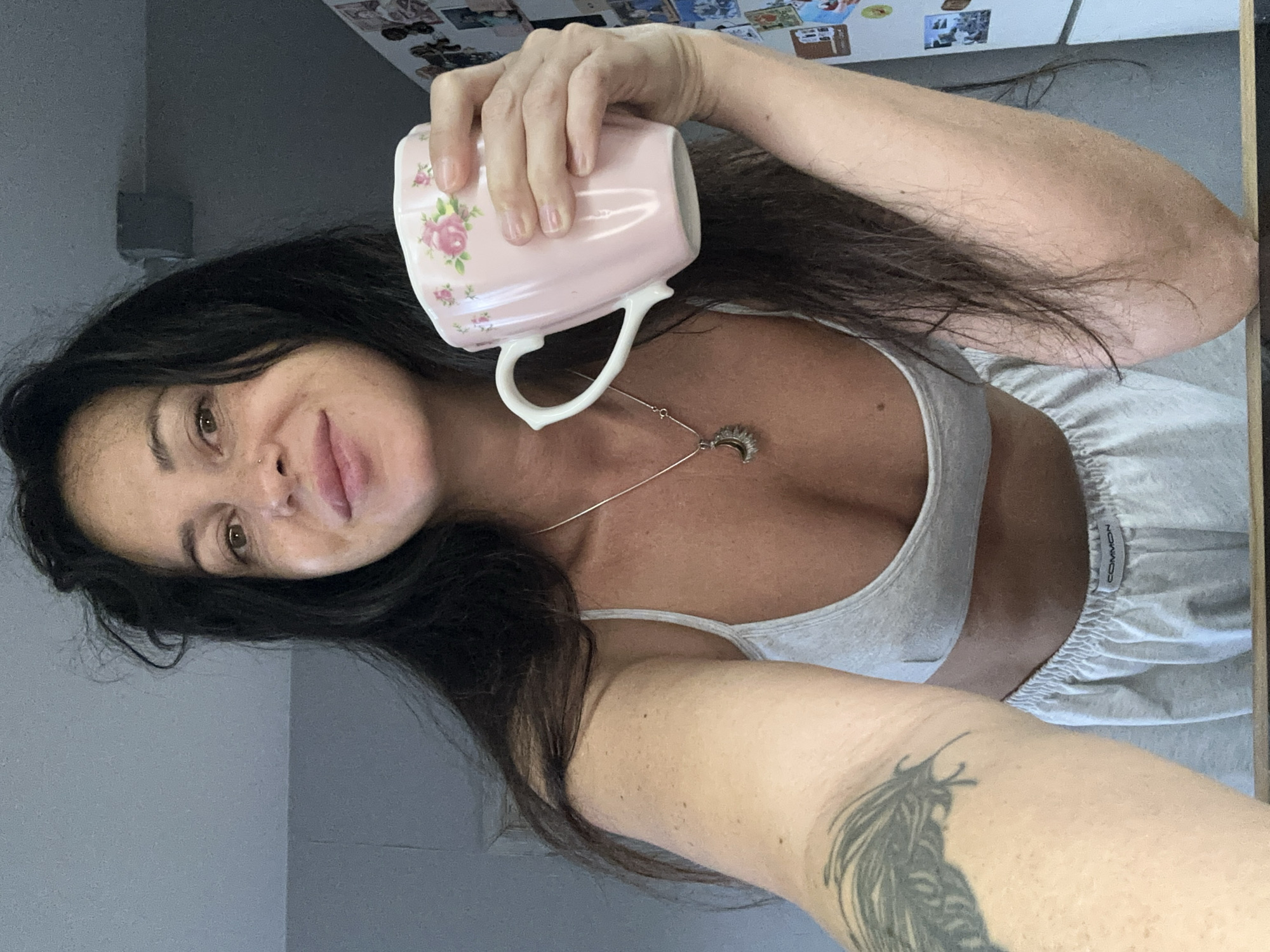 The image shows a woman indoors, holding a pink mug with a floral pattern. She is wearing a grey sports bra and grey sweatpants. Her dark hair is long and appears slightly tousled. A silver necklace with a crescent moon pendant is visible around her neck. A tattoo of a feather is present on her left arm. The background suggests an indoor domestic setting, possibly a kitchen, with a refrigerator visible on the right side adorned with magnets or pictures. The overall lighting is bright and natural, indicating daytime. There is no information available about the specific location in the city of Irkutsk, Russia.