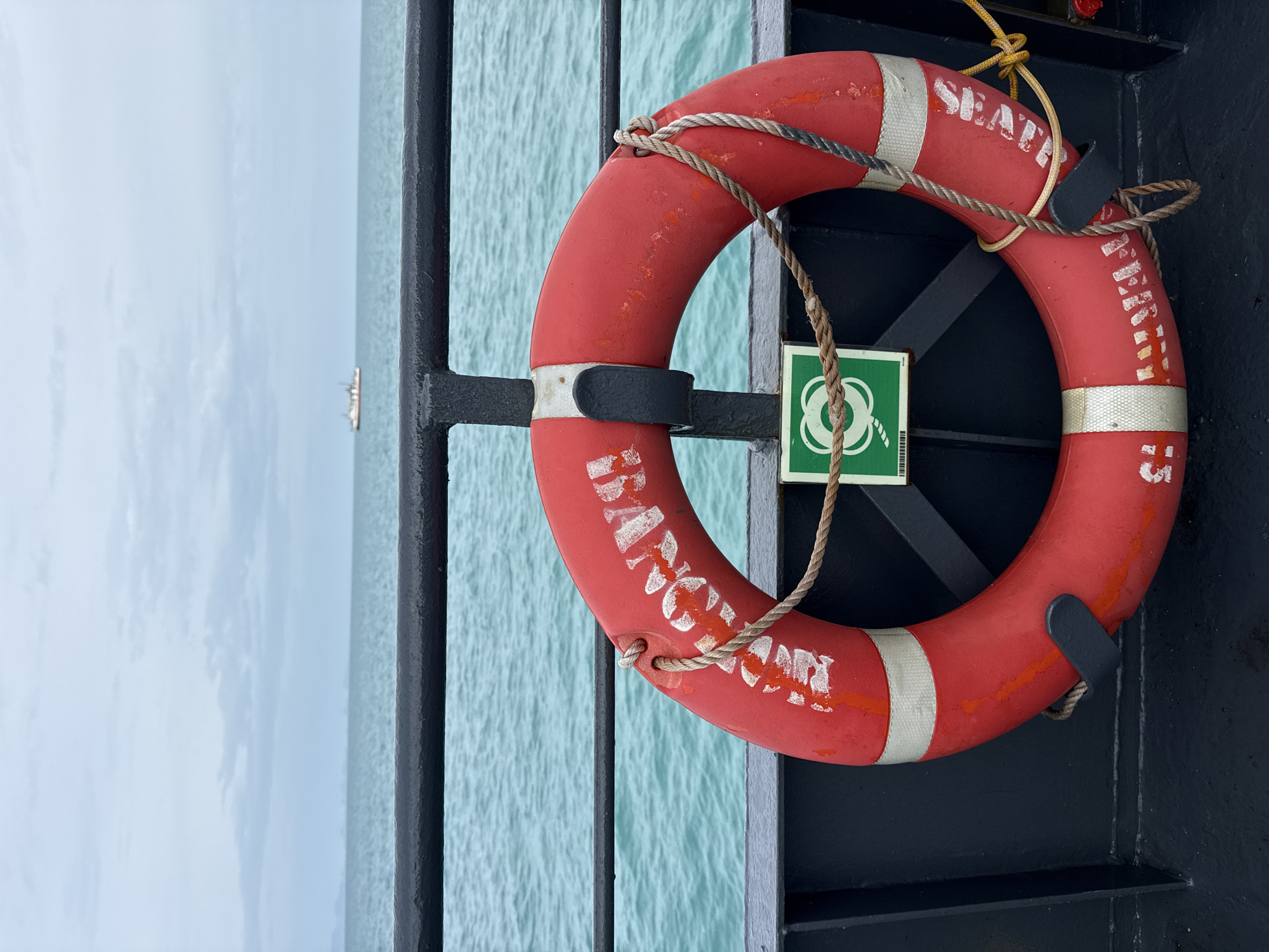 A red life preserver is attached to a dark railing on the deck of a ferry, with the turquoise water of the Gulf of Thailand visible beyond the railing. The life preserver has white lettering that reads "BANGKOK" and a green sign with a white life preserver symbol is attached to the railing below it. In the distance, a small white boat with an orange stripe is visible on the horizon. The sky is overcast. The scene appears to be on a ferry traveling near Ko Samui District, Thailand.