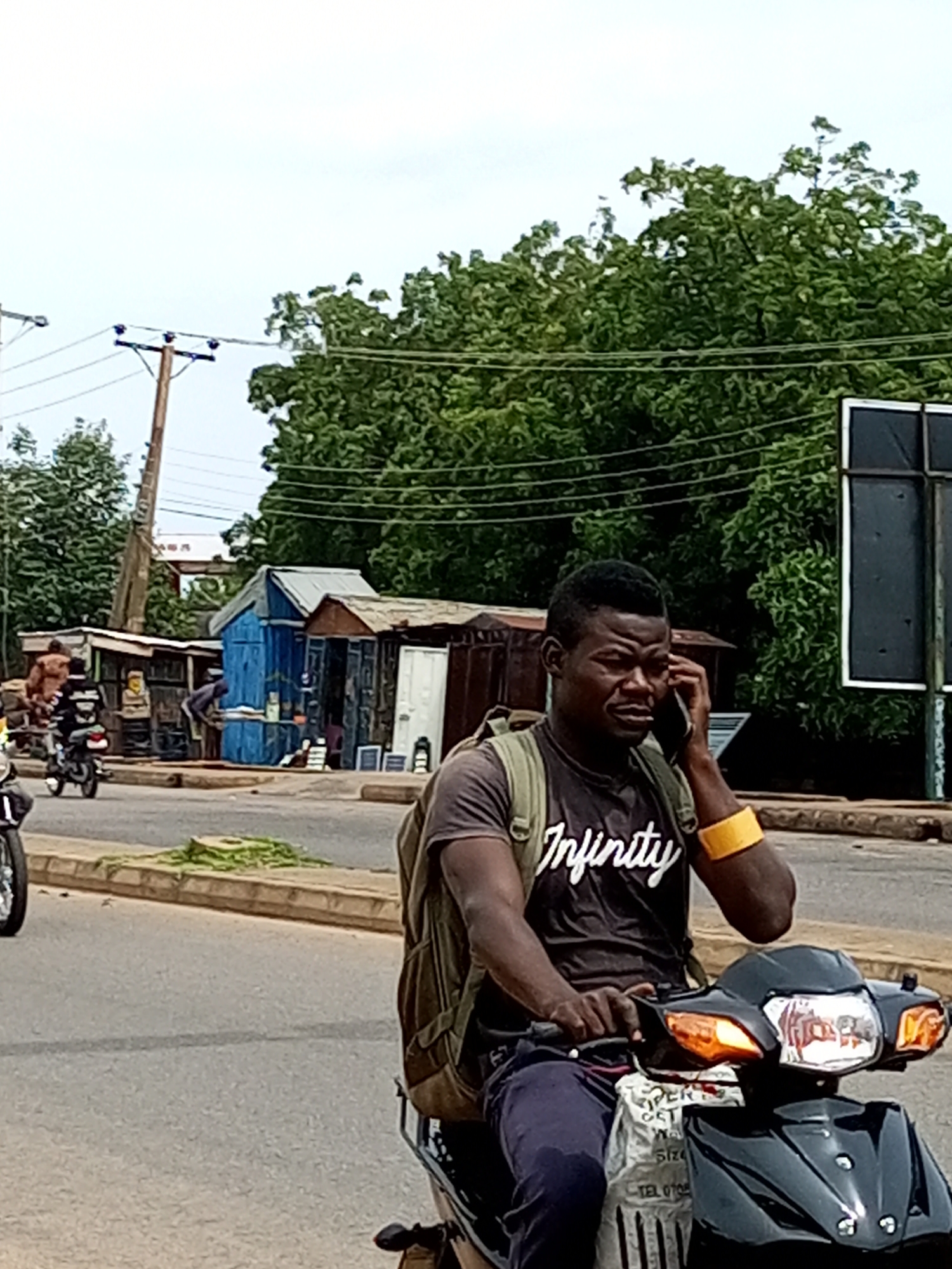In the city of Garko, Nigeria, a man wearing a dark t-shirt with the word "Infinity" printed on it and a backpack is seen riding a black motorcycle. He is holding a cell phone to his right ear and appears to be in conversation. A yellow band is visible on his left wrist. The motorcycle has orange turn signals and a white bag hanging from the front. In the background, several market stalls with corrugated metal roofs are visible, along with trees and power lines. Other people and motorcycles can be seen in the distance. The scene depicts a typical street view with commerce and transportation.