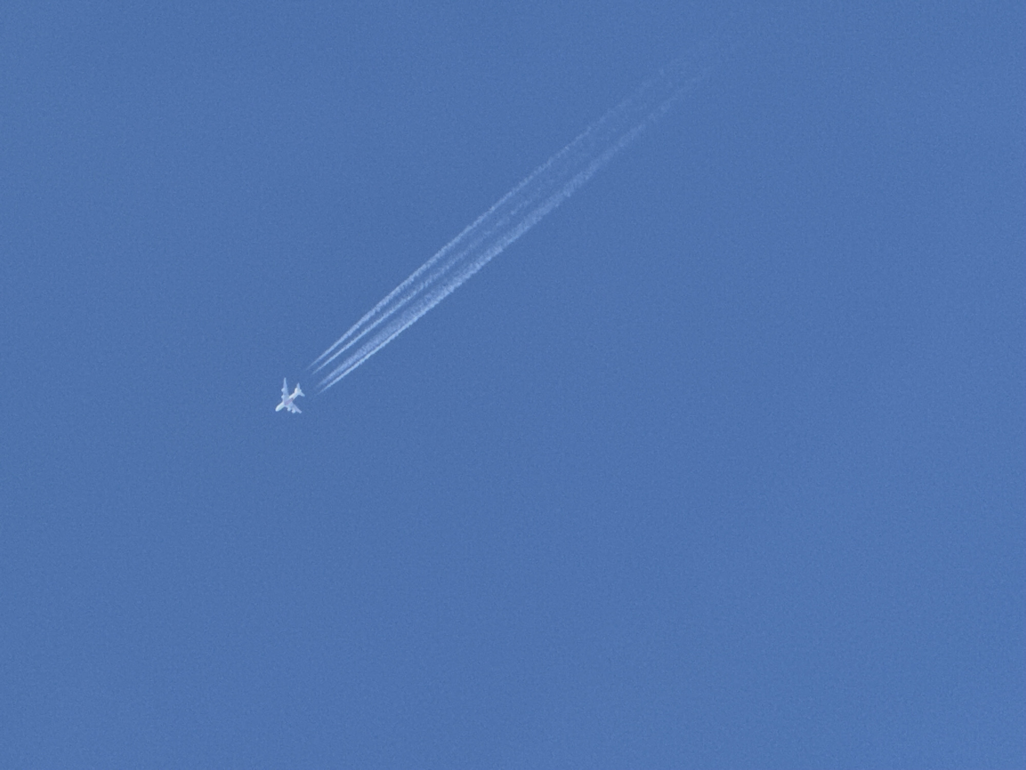 The image shows a clear blue sky with a small airplane leaving a contrail. The airplane is white and appears to be a jet. The contrail is long and trails behind the plane, spreading out in a wispy fashion. The sky is a uniform shade of blue, indicating good weather and likely daytime. The perspective is from below, looking up at the airplane. There is no visible text in the image. The overall scene is peaceful and evokes a sense of vastness and travel. The photograph is likely taken in an outdoor setting, possibly during the day due to the bright sky. The focus is on the airplane and its contrail, highlighting the solitary nature of the aircraft against the expansive sky.