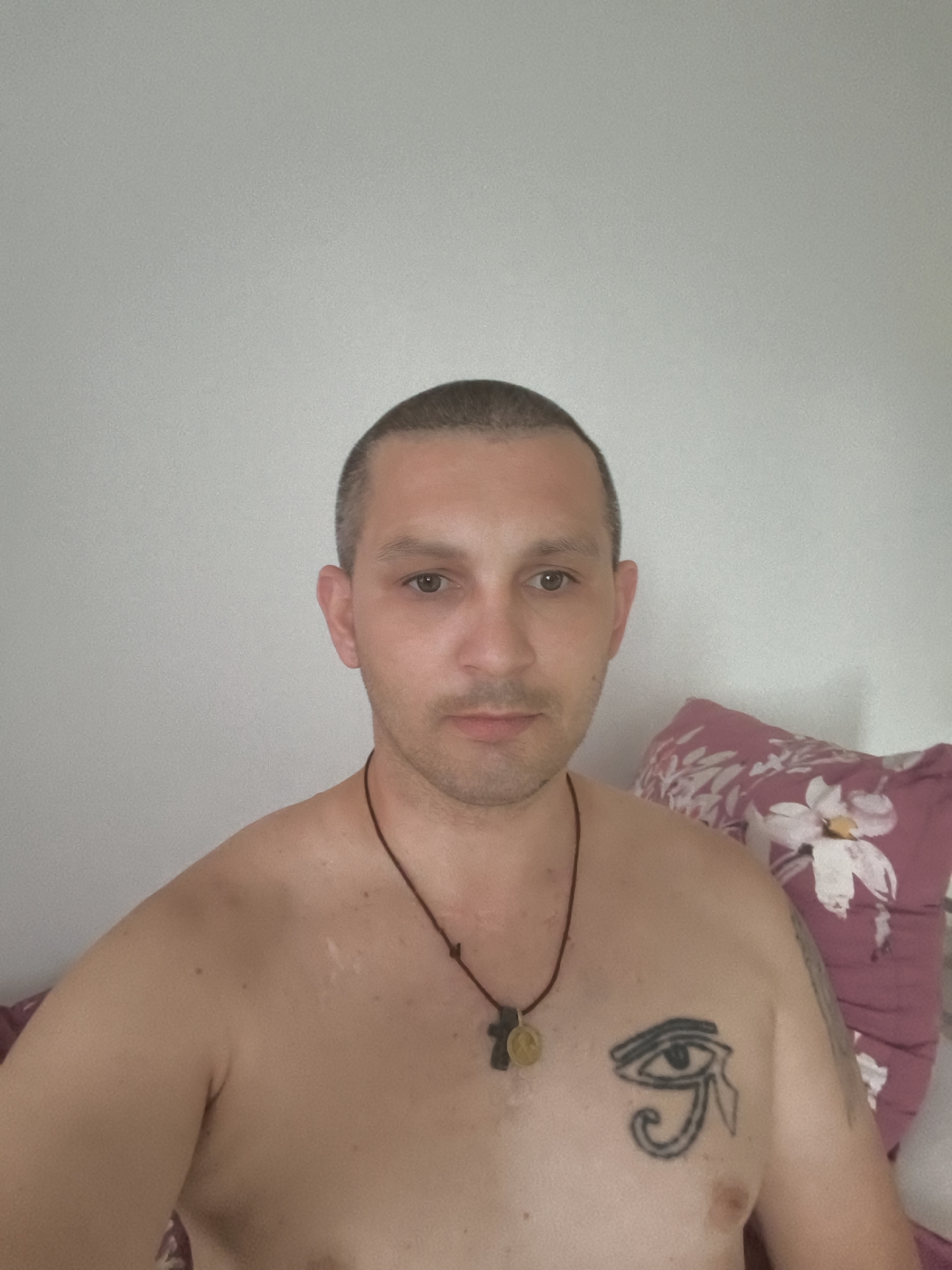 A shirtless man with a buzz cut and an Egyptian eye tattoo on his chest is posing for a selfie in a bedroom. He is wearing a dark necklace with two pendants, one of which appears to be a cross. Behind him, a floral pillow rests against a plain wall. The man is looking directly at the camera with a neutral expression. The overall lighting suggests it is daytime, and the setting appears to be indoors. There is no information in the image to suggest the location in the city of Wolgast, Germany.