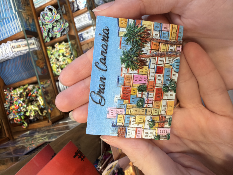 The image displays a rectangular souvenir magnet, approximately 10-12 cm in width, held horizontally by a person's light-skinned hands in an indoor retail setting. The magnet features the embossed dark brown text "Gran Canaria" in a cursive font, set against a light blue background representing the sky. Below the text, a vibrant, three-dimensional relief depicts a dense, colorful townscape with numerous multi-story buildings in varying hues including pink, yellow, blue, red, orange, and white, suggesting a hillside settlement. Three palm trees are interspersed within this townscape, with two prominent ones on the right side. A small blue dome structure is visible at the bottom center of the magnet's illustration. A gold-colored ring is present on the ring finger of the left hand. The background, out of focus, reveals wooden display shelving filled with assorted souvenir merchandise, including small colorful figurines and packaged items. The scene is consistent with a souvenir shop located in the city of Arucas, Gran Canaria, Spain.