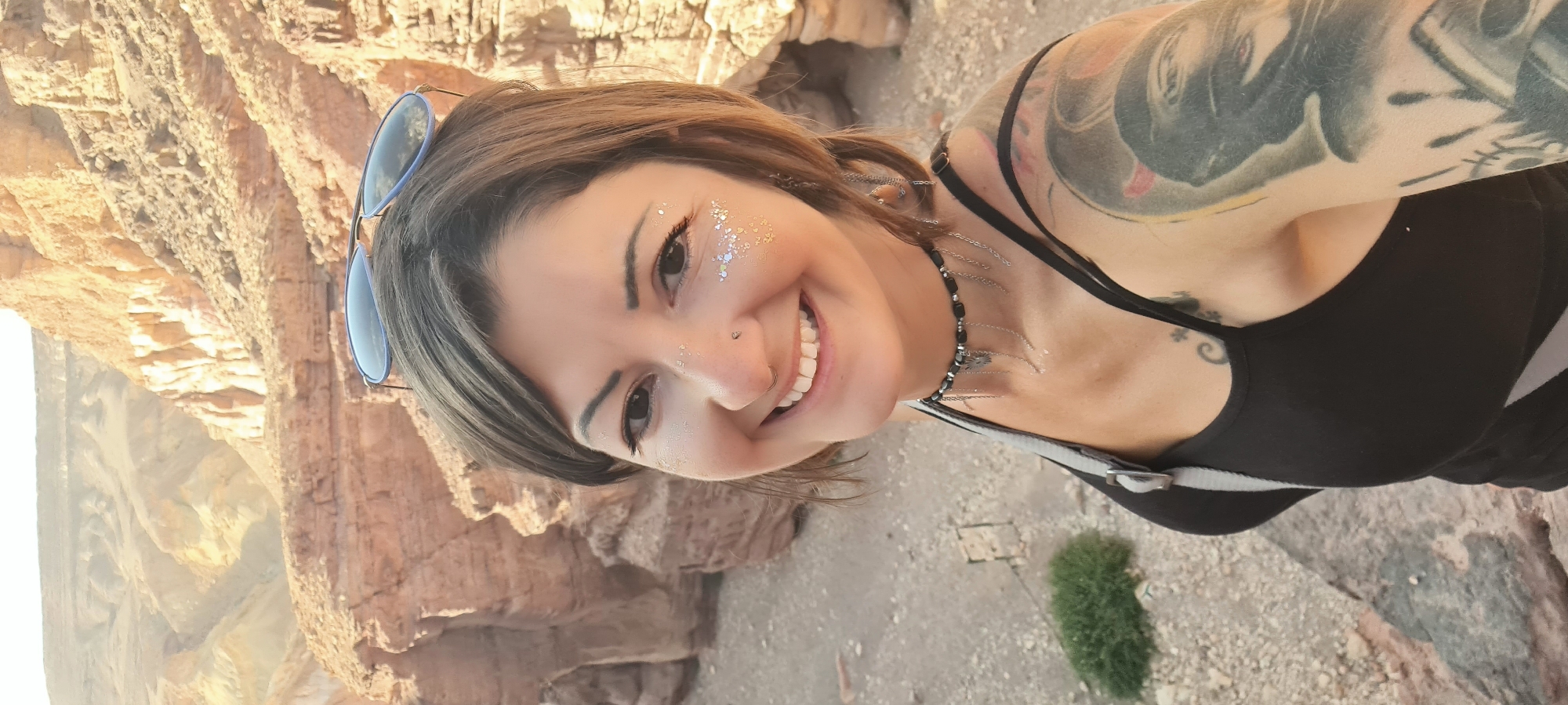 A woman with tattoos on her arm and neck is smiling at the camera. She has glitter makeup around her eyes, a nose ring, and sunglasses on her head. She's wearing a black top and a necklace. The background shows a rocky landscape. The location is identified as Hevel Eilot Regional Council, Israel.