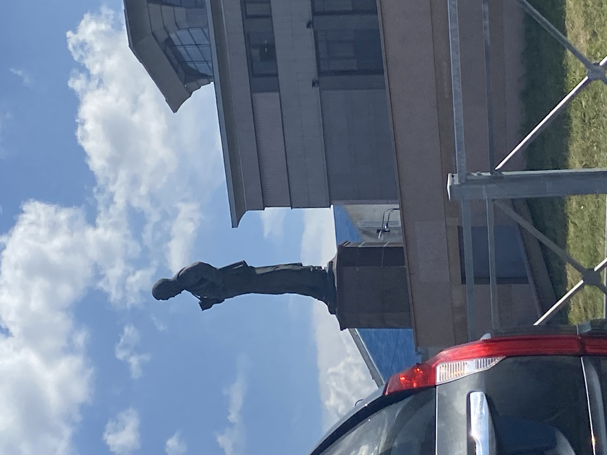 The image depicts a statue of a man in profile, standing on a pedestal against a bright blue sky with scattered white clouds. The statue is a dark silhouette, suggesting it is made of a dark material or is in shadow. The man is wearing formal attire, possibly a suit, and is holding an object in his hands near his chest. To the right of the statue, a modern building with large windows and a distinctive angled roofline is visible. The building appears to be made of concrete and glass. In the foreground, a portion of a dark-colored vehicle is visible on the left, with its red taillight illuminated. A metal fence with a diagonal crossbar runs across the lower portion of the image, separating the foreground from a grassy area behind it. The overall scene suggests an outdoor urban environment. Based on the provided crops and external information, this location is identified as the monument to Musa Cälil in Kazan, Russia.