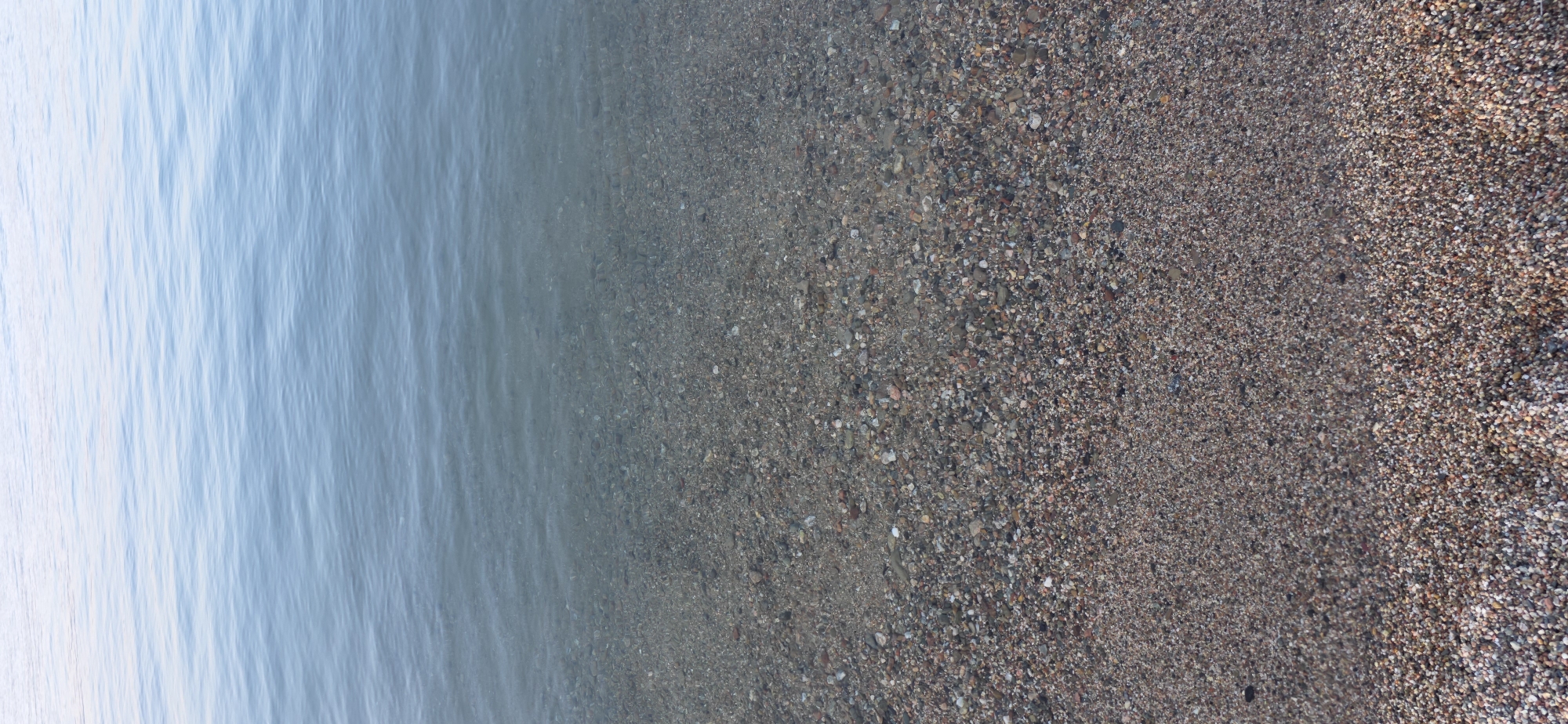 This image depicts a tranquil scene at the shore of a body of water, likely a lake or calm sea. The water occupies the upper portion of the frame, exhibiting gentle ripples and a gradient of light blue hues that suggest clear, shallow depths. The bottom half of the image showcases a beach composed of fine pebbles and sand, transitioning from wet, darker stones submerged at the water's edge to drier, lighter colored material further inland. The overall impression is one of natural serenity, devoid of any human presence or artificial elements. The location is identified as Boz Beshik, Kyrgyzstan.