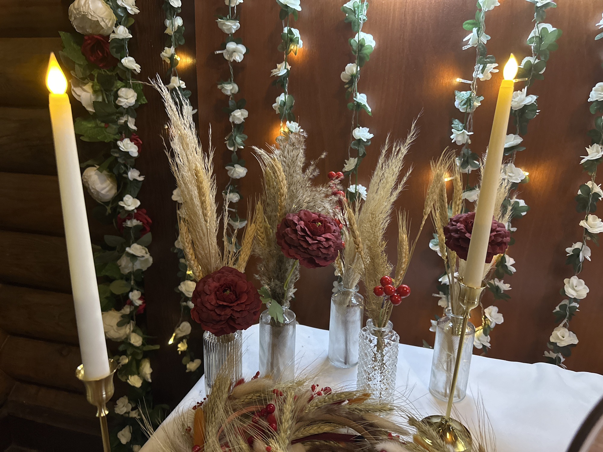 A still life arrangement is displayed on a white tablecloth, set against a warm brown wooden background. The background is adorned with two vertical garlands of artificial white flowers and green leaves, intertwined with small, warm-toned fairy lights.
Dominating the foreground on the left is a tall, white, LED candle in a brass candlestick, its flame glowing. To the right of the candle, a series of glass vases are arranged. These vases contain dried wheat stalks, some with a reddish-brown hue, and prominent, deep red, peony-like flowers. One vase also holds a small cluster of bright red berries.
In the lower center of the image, a circular arrangement of dried wheat stalks and grasses, interspersed with red berries and white fluffy elements, lies on the tablecloth.
The overall impression is one of rustic elegance, possibly for a special occasion or decorative display. There are no people or animals present in the image. The specific location within Novokuznetsk, Russia, cannot be determined from the visual information provided.